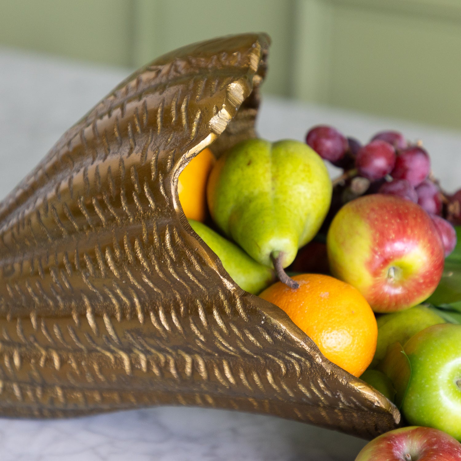 A decorative metallic gold cornucopia-style fruit bowl filled with oranges, pears, apples, and purple grapes on a light marble surface.