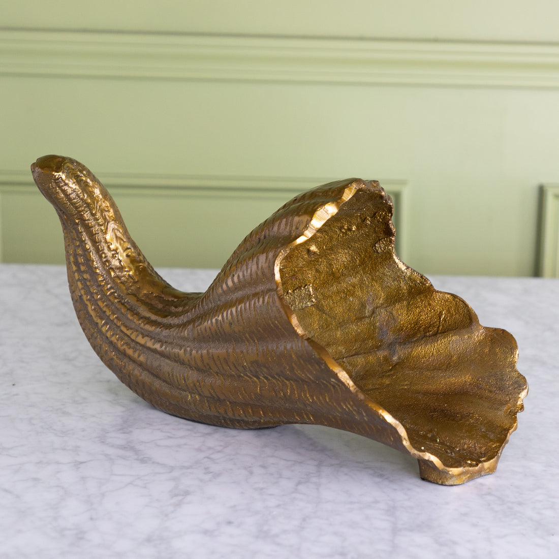 A decorative gold-toned horn-shaped bowl with a textured, ribbed surface and an irregular, scalloped rim, displayed on a white marble surface against a pale green paneled wall.