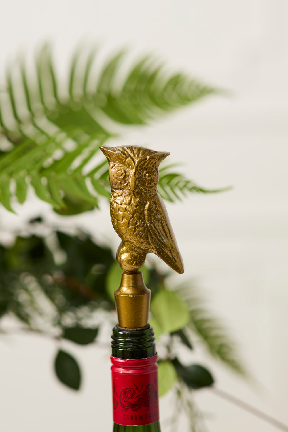 Gold owl-shaped bottle stopper on a red-wrapped wine bottle with a blurred green plant background.