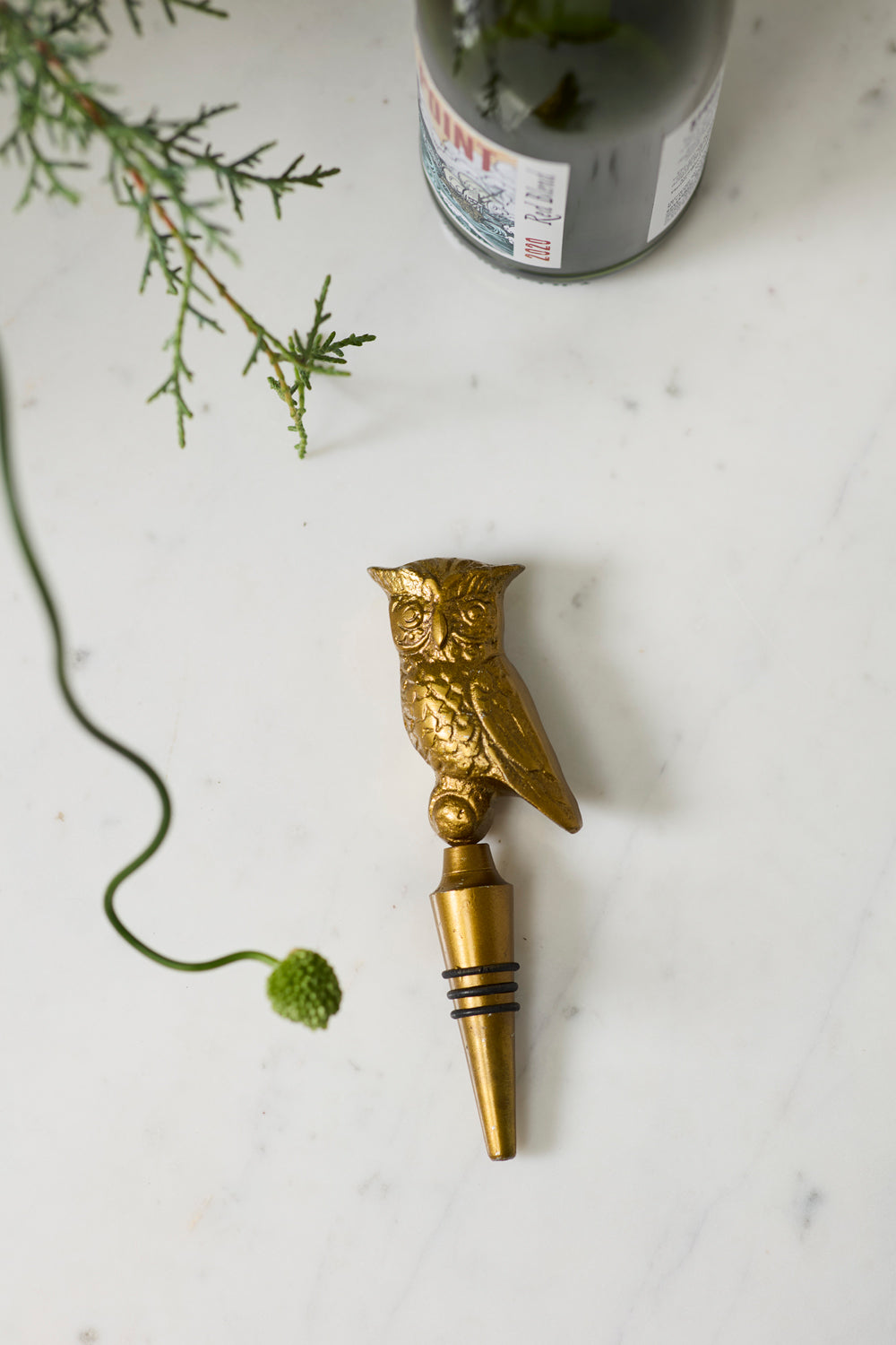 Gold owl-shaped bottle stopper on a light surface with greenery.