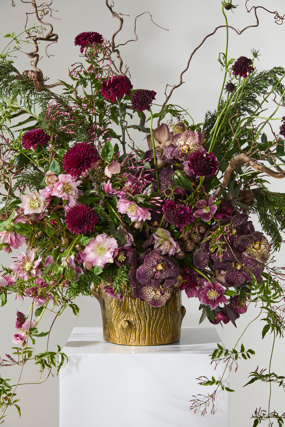 Floral arrangement with purple and pink flowers in a gold vase on a white surface.