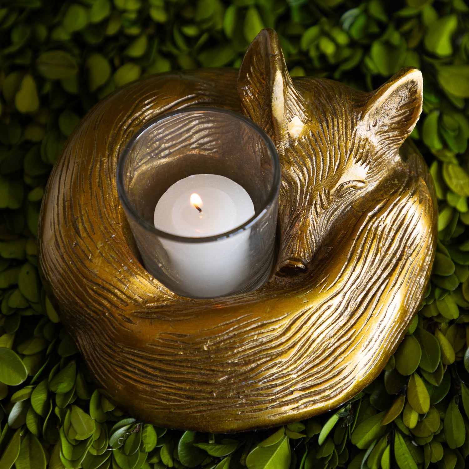 Gold fox-shaped candle holder with a lit white candle on a green leafy background.
