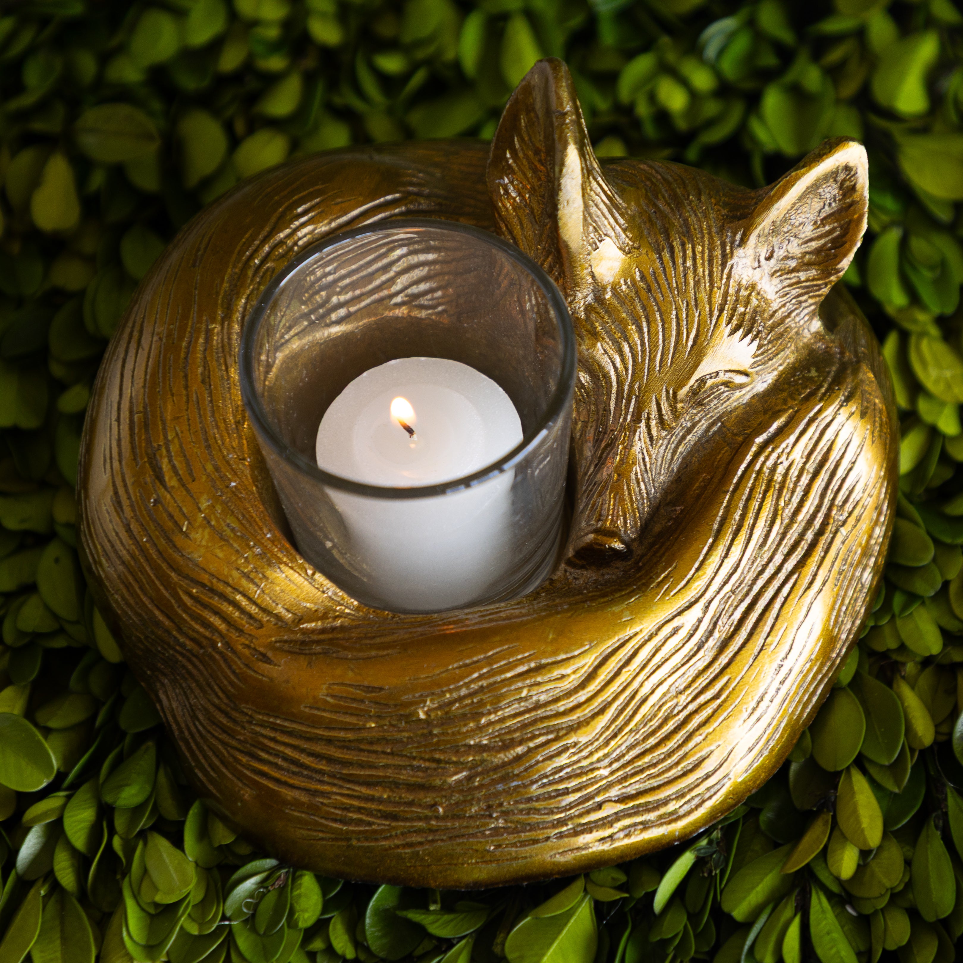 Gold fox-shaped candle holder with a lit white candle on a green leafy background.