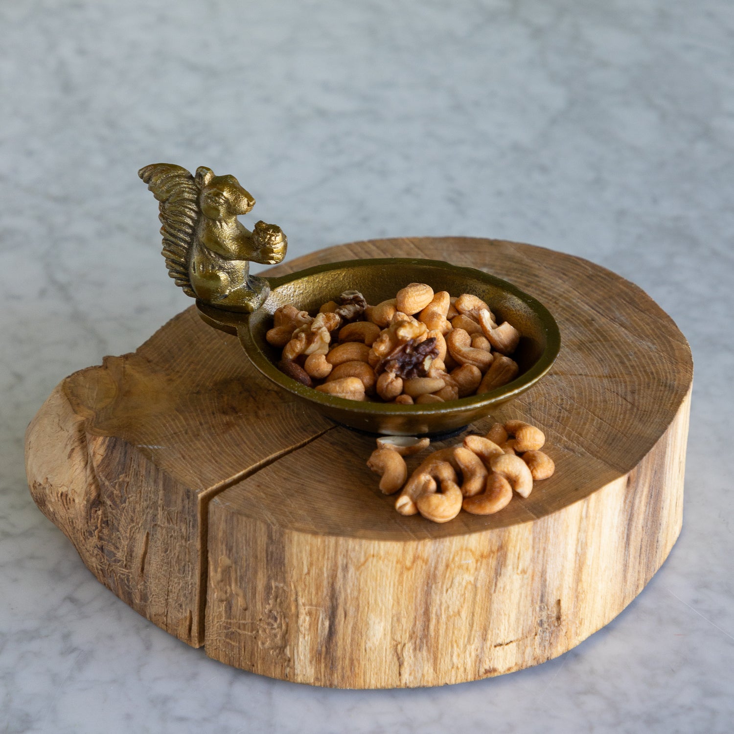 A small brass bowl with a sculpted squirrel handle, filled with cashews and walnuts, displayed on a rustic round wood slice stand against a light gray marble background.