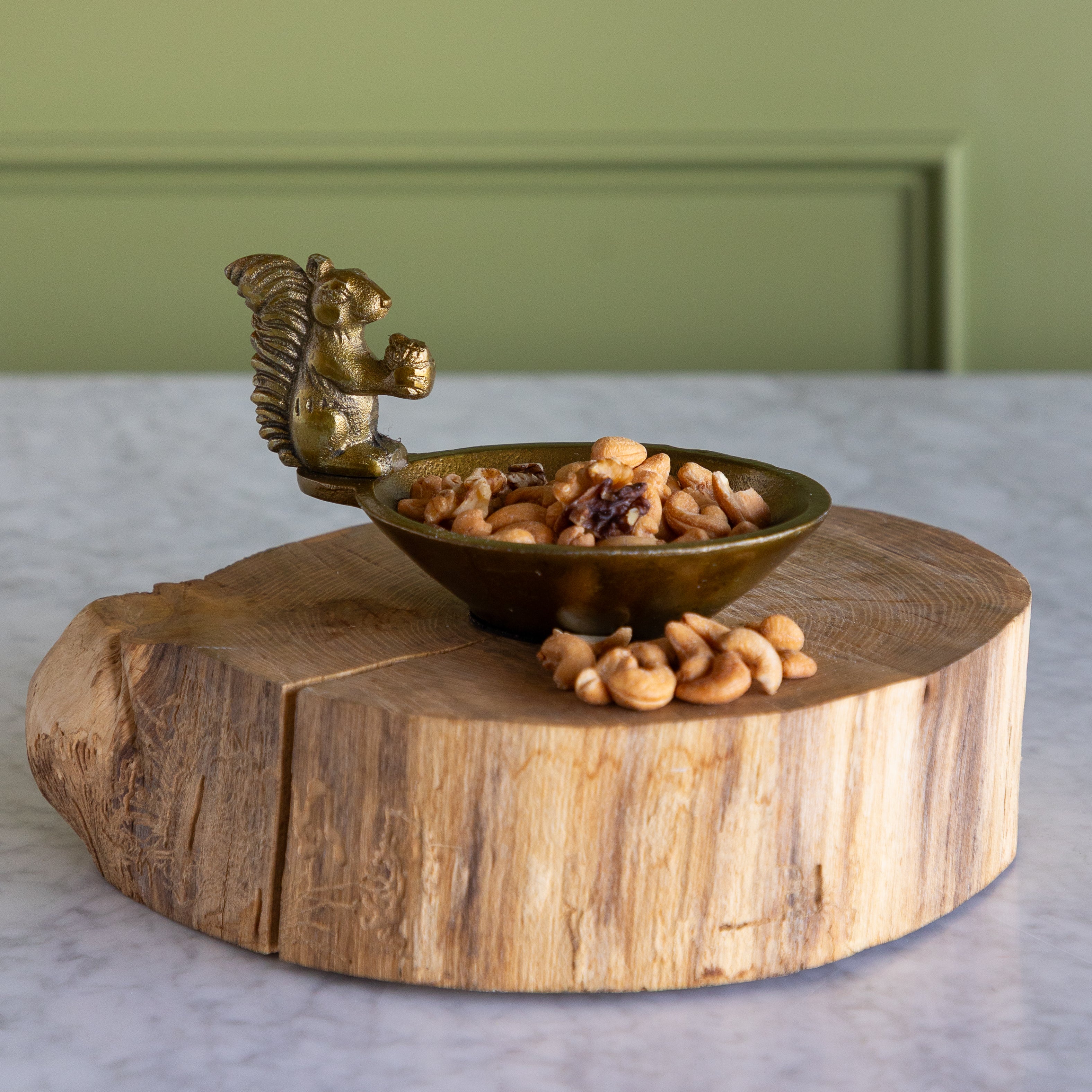 A decorative bowl with a squirrel-shaped handle, filled with cashews and dried fruit pieces, displayed on a rustic round wooden slice stand against a green paneled wall and light marble surface.