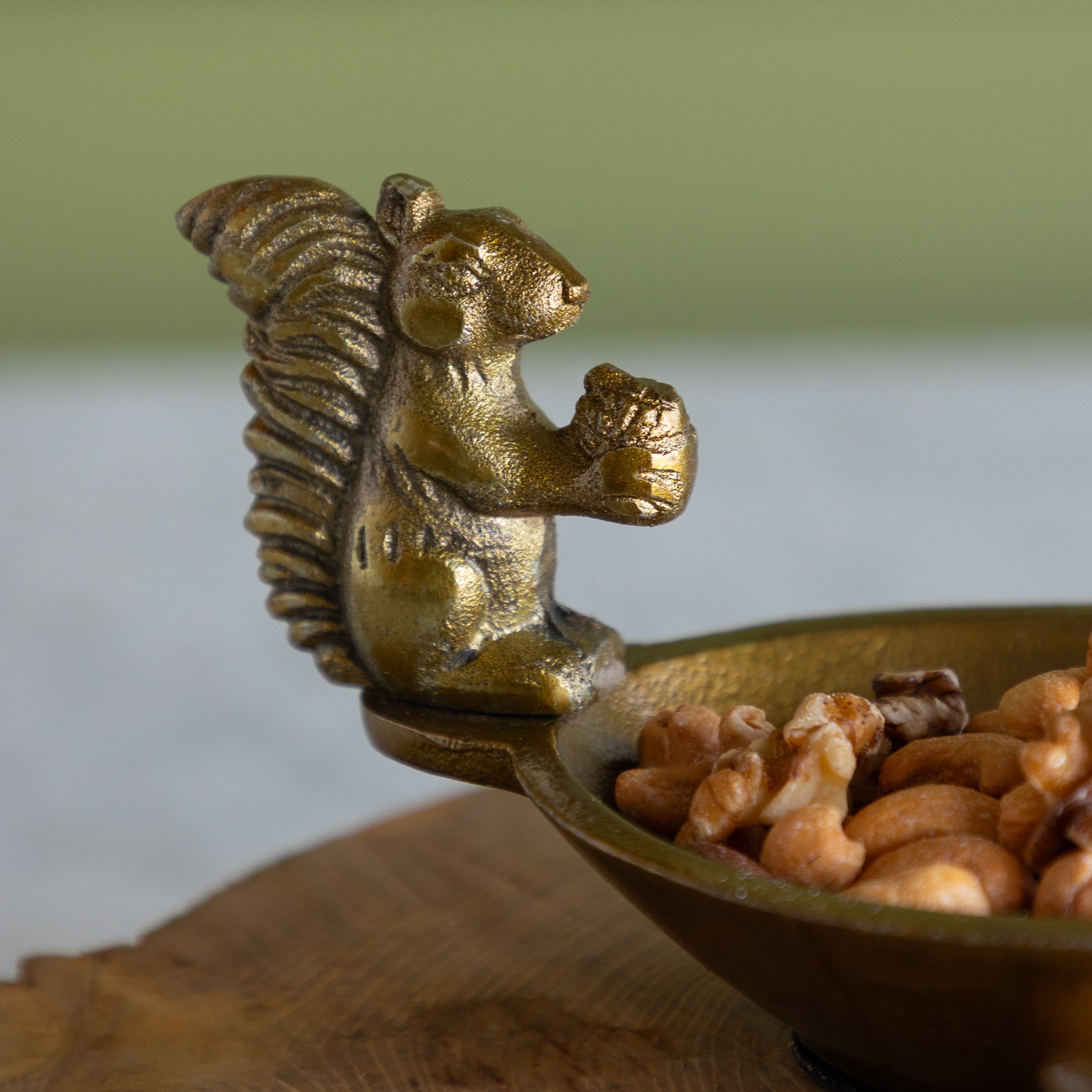 A close-up of a decorative brass squirrel-shaped bowl handle holding walnuts in a bowl, set on a wooden surface with a soft-focus green and gray background.