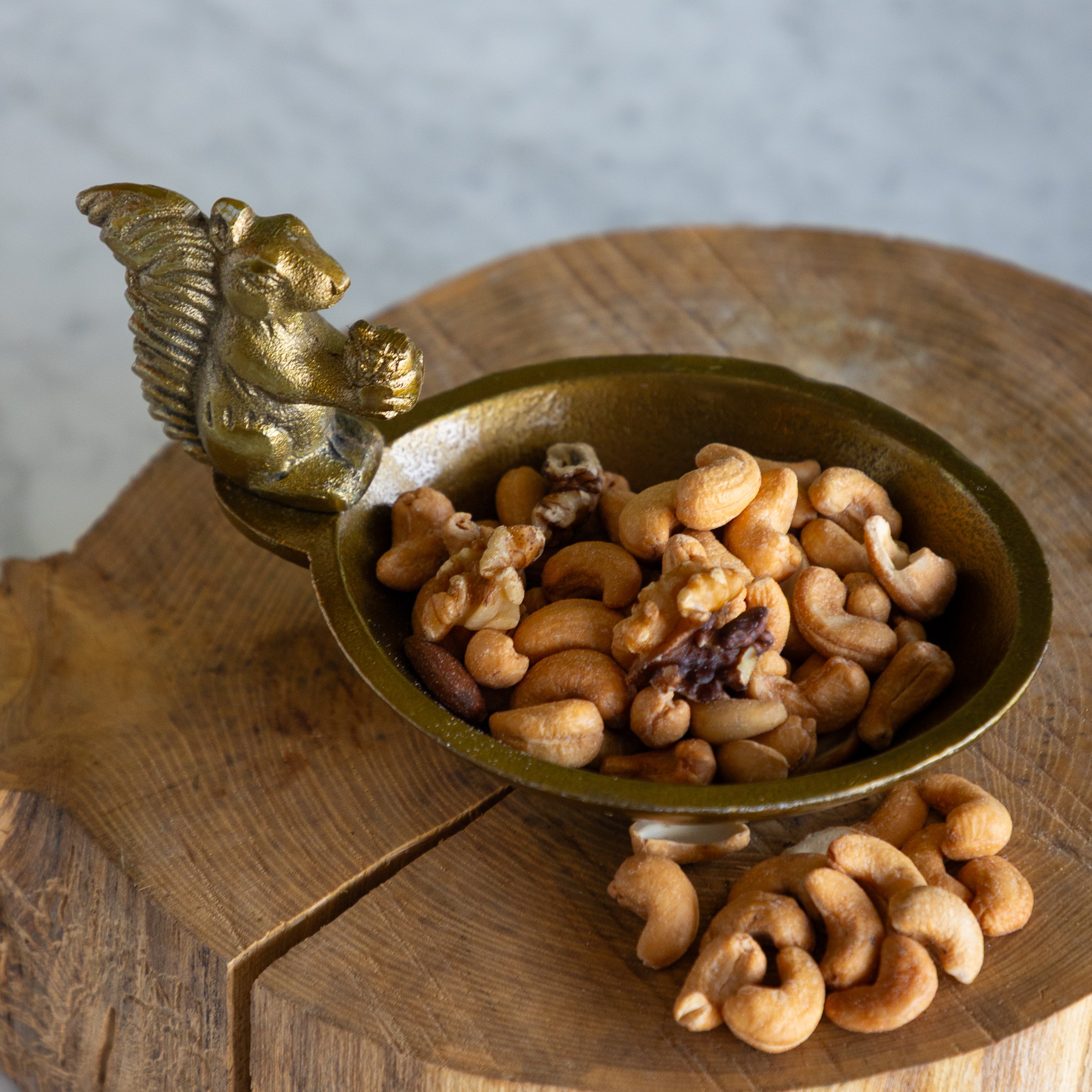 A small ornate metallic bowl with a sculpted handle, filled with mixed nuts including cashews and walnuts, set on a round wooden board against a light gray background.