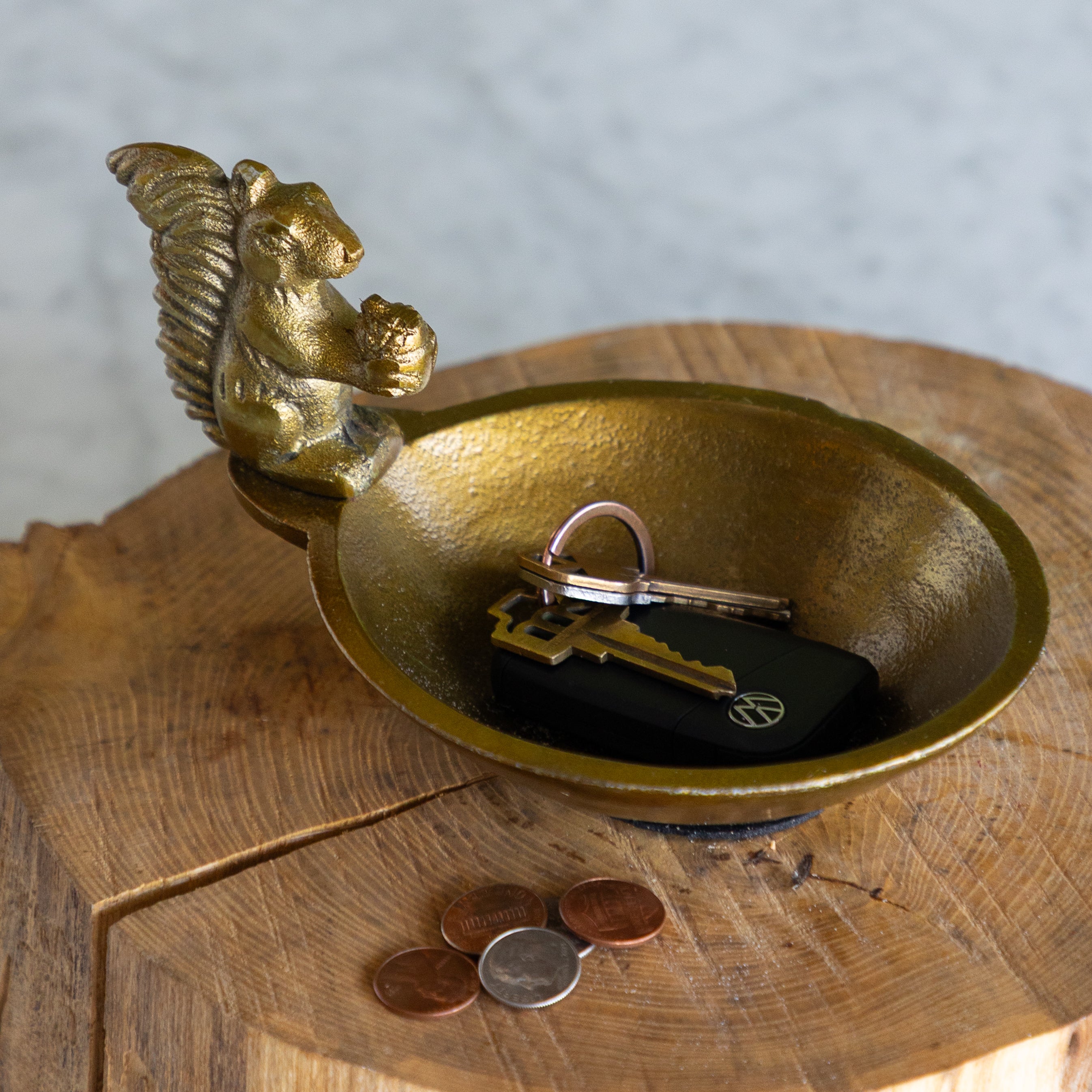 A brass key holder bowl with a brass squirrel perched on the rim, shown holding keys and a black car key fob on a rustic wooden surface.