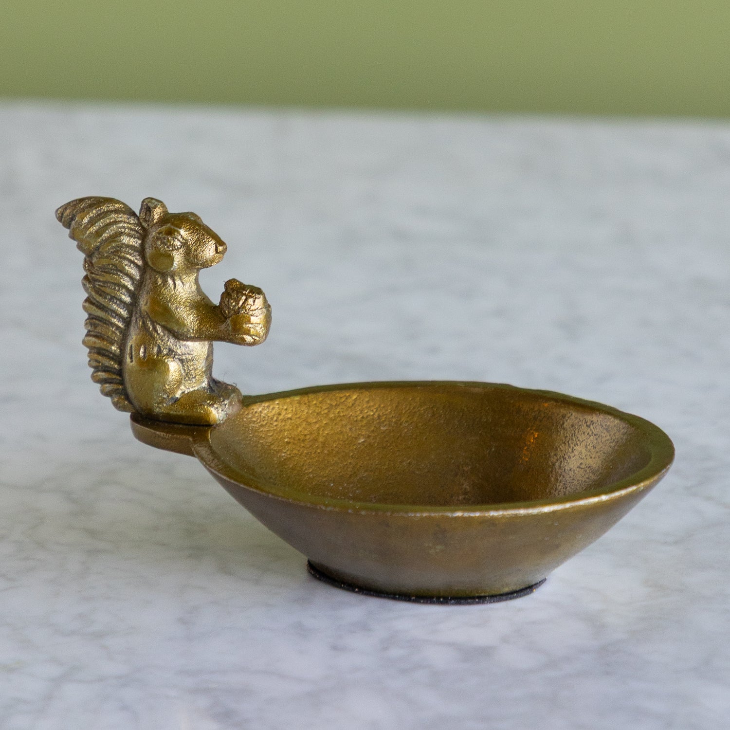 A small metallic bowl with a sculpted squirrel handle, shown on a light marble-like surface with a soft green background.