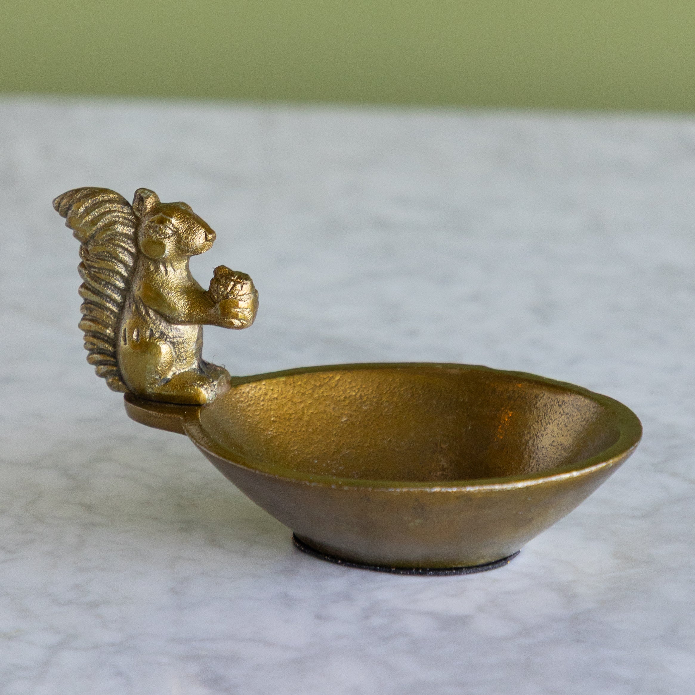A small metallic bowl with a sculpted squirrel handle, shown on a light marble-like surface with a soft green background.