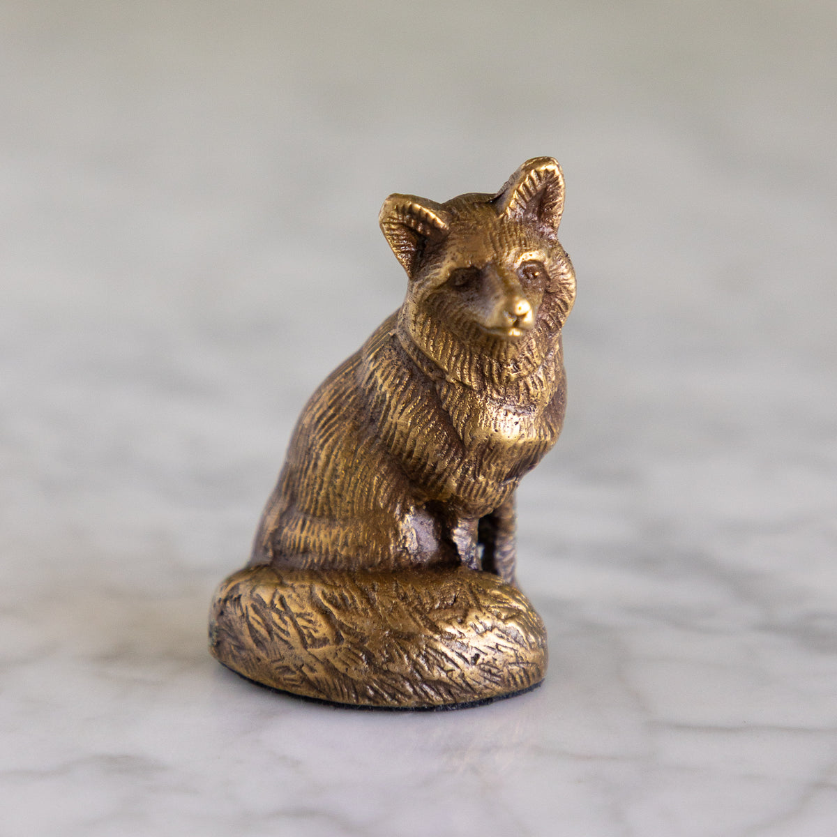 A small metallic fox figurine in a warm gold/bronze finish, sitting on a round textured base against a soft gray marble-like background.