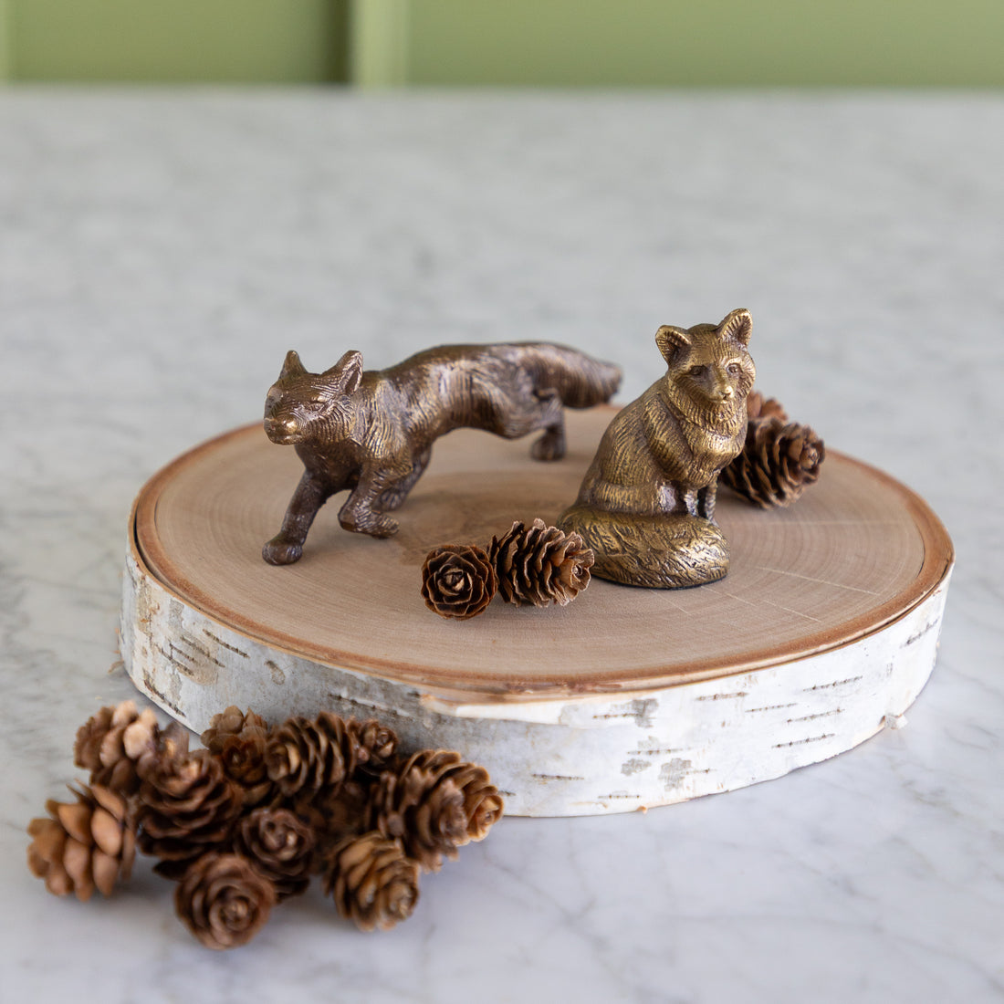 Two small metallic animal figurines, one fox and one wolf, displayed on a round birch log slice with pinecones on a light marble surface.