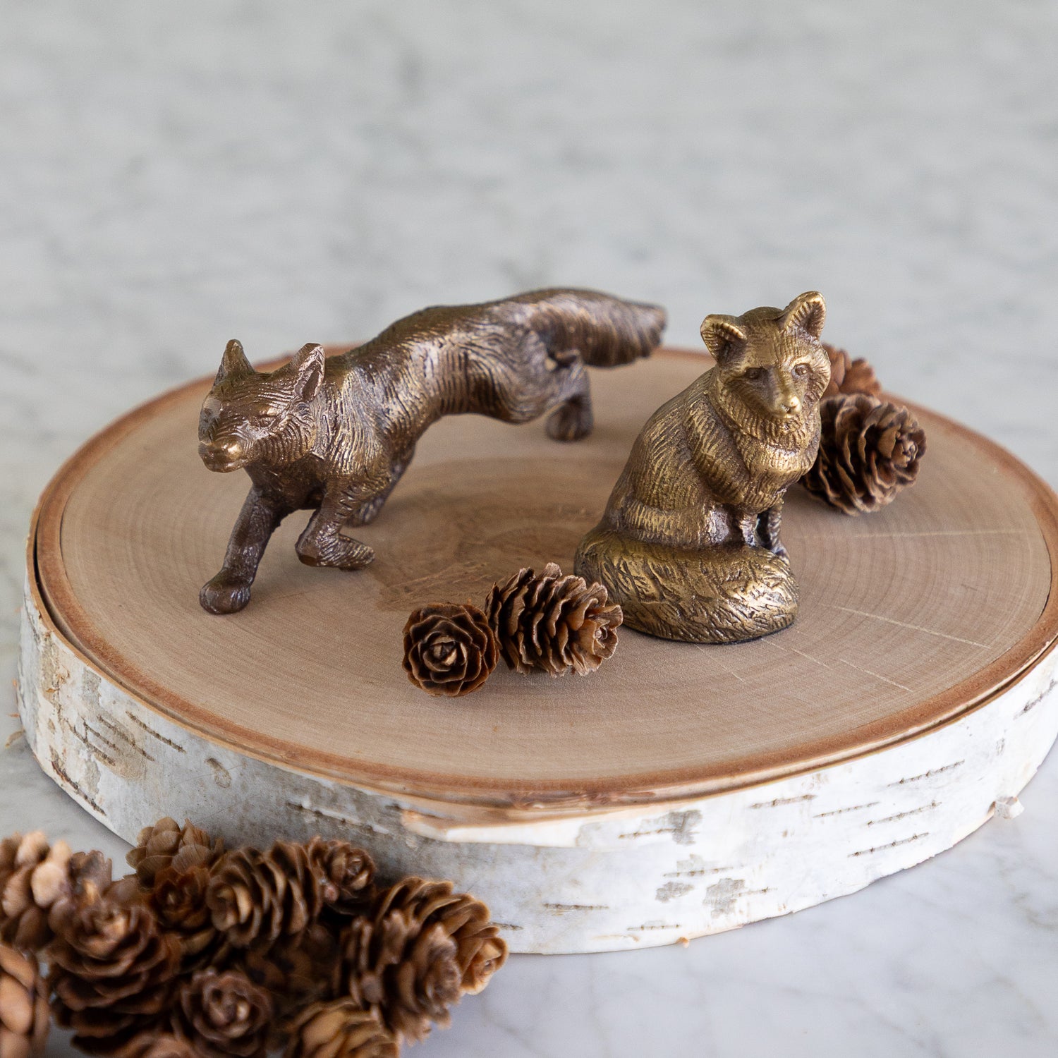Two small metallic animal figurines, one fox standing and one cat sitting, displayed on a round birch wood slice with pinecones on a light marble surface.