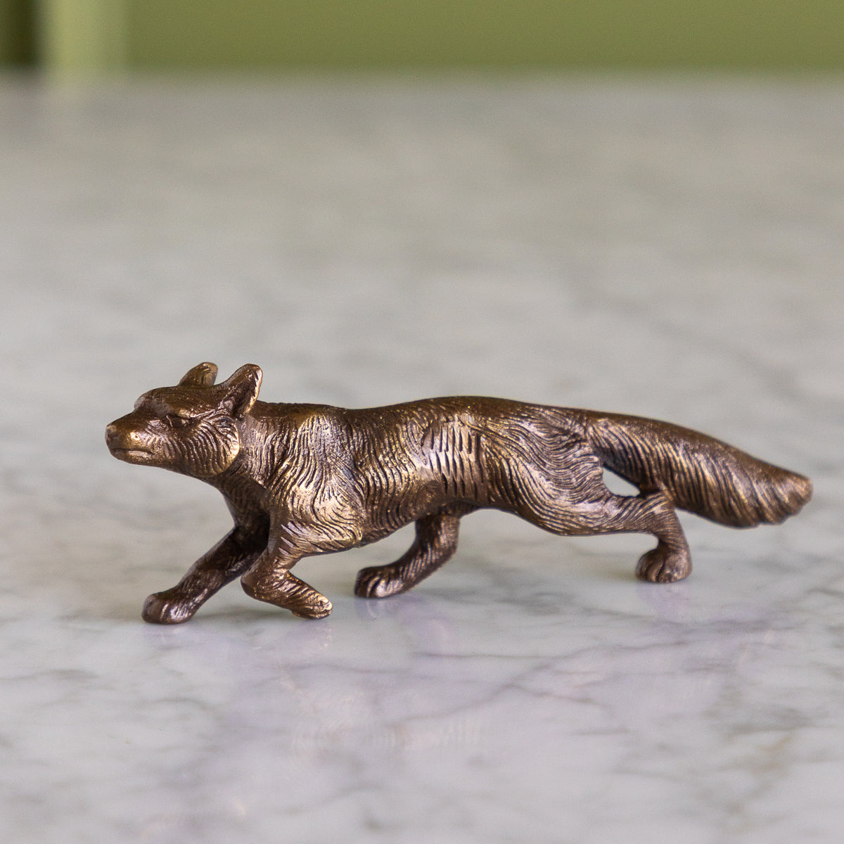 A small metallic fox figurine in a bronze finish, shown in profile on a light marble surface with a softly blurred green background.