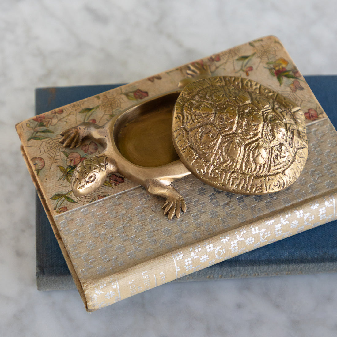 A brass turtle-shaped keepsake box with an embossed lid, resting on top of vintage floral books on a marble surface.
