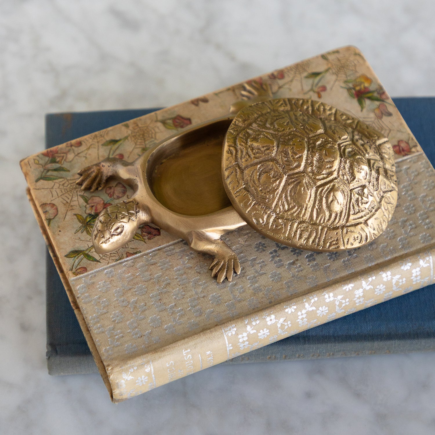 A brass turtle-shaped keepsake box with an embossed lid, resting on top of vintage floral books on a marble surface.