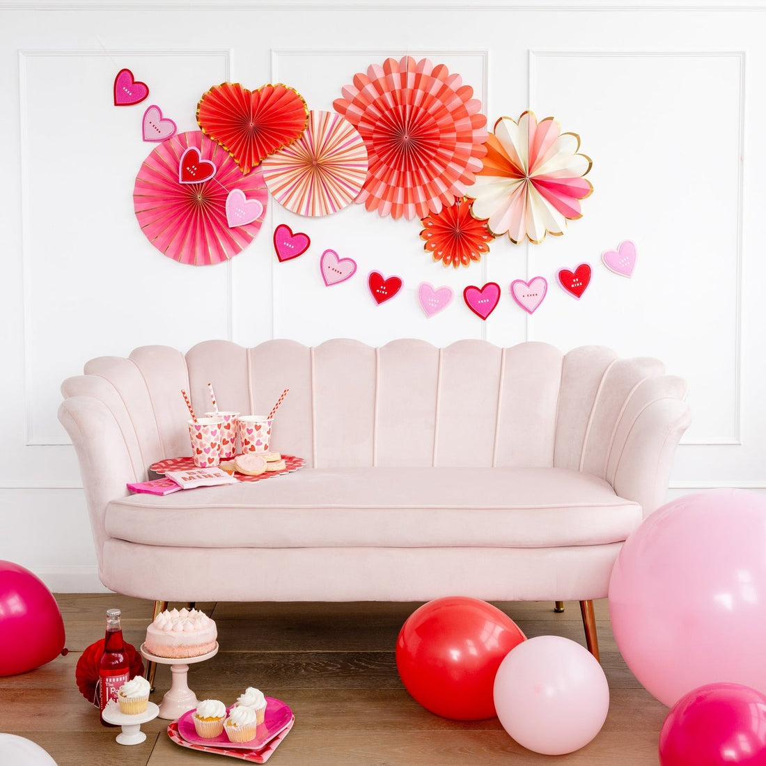 A party setting with a couch and set of 6 different shaped paper fans in assorted shades of pinks with gold accents on the wall above a couch. 