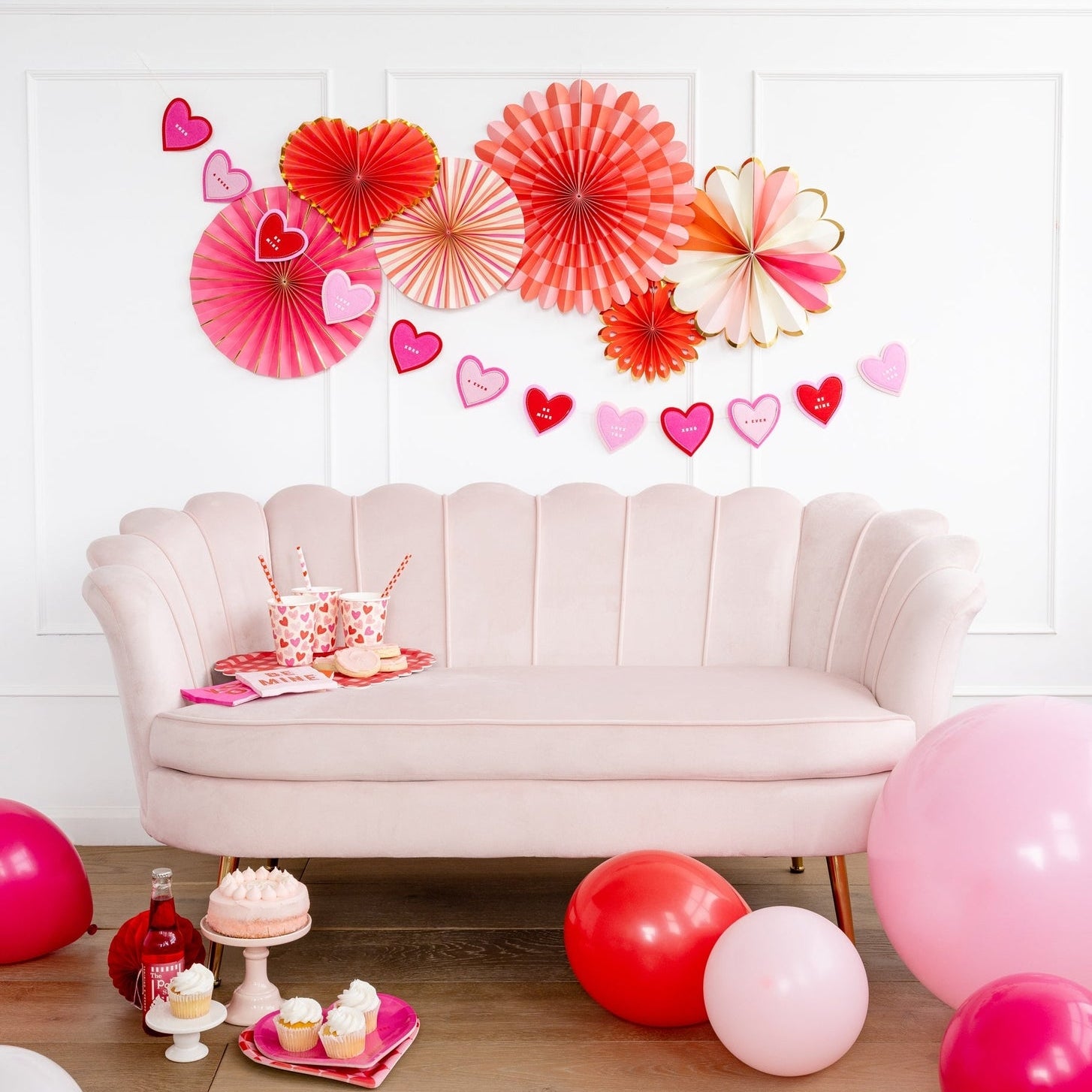 A party setting with a couch and set of 6 different shaped paper fans in assorted shades of pinks with gold accents on the wall above a couch. 
