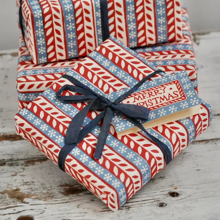 Stack of Christmas-themed wrapped presents on a wooden surface with a Merry Christmas card tucked into the ribbon. 