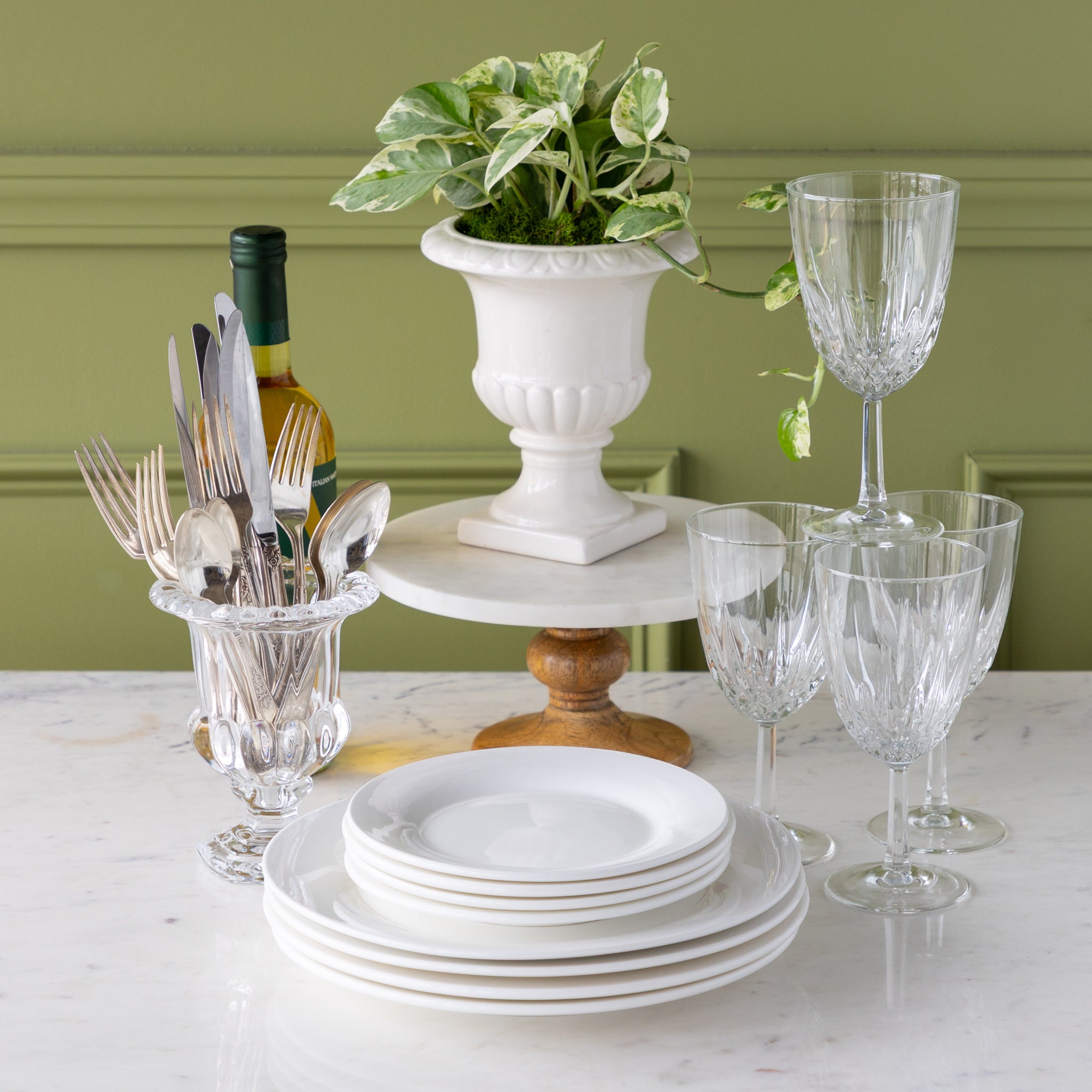 Dining table setting with plates, glasses, cutlery, and a plant on a white surface.
