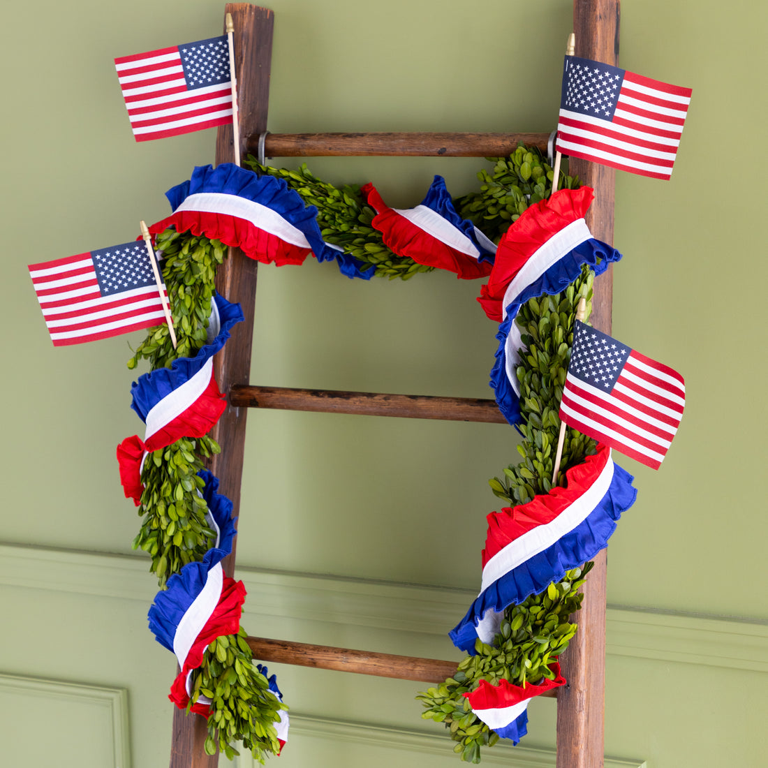 Decorative ladder with American flags and ruffled patriotic ribbon wrapped around greenery against a green wall.