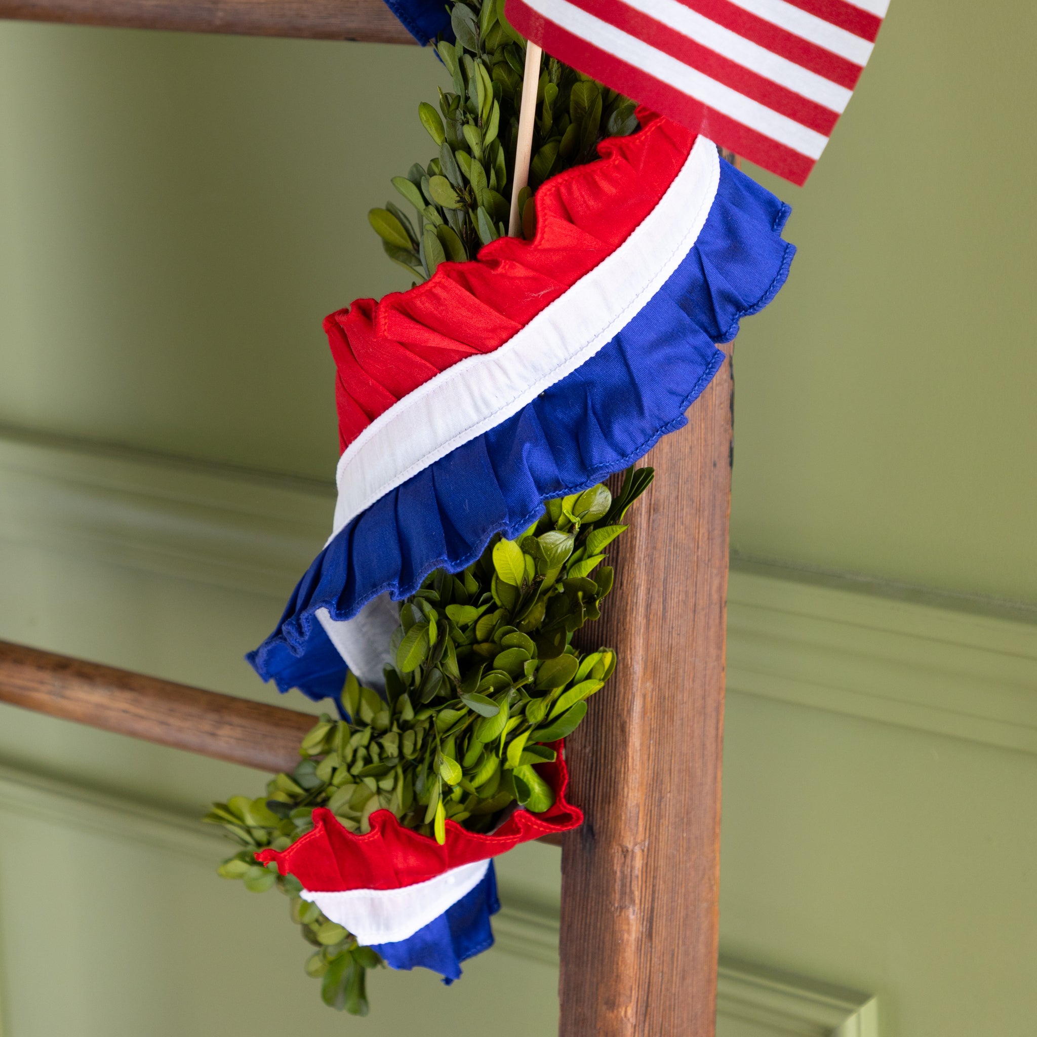Decorative ladder with American flags and ruffled patriotic ribbon wrapped around greenery against a green wall.