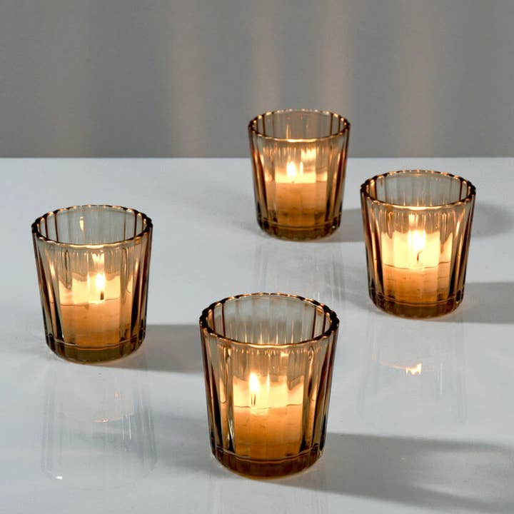Four amber glass candle holders with a ribbed fluted design, each containing a lit tealight candle, arranged on a white reflective surface against a neutral gray background.