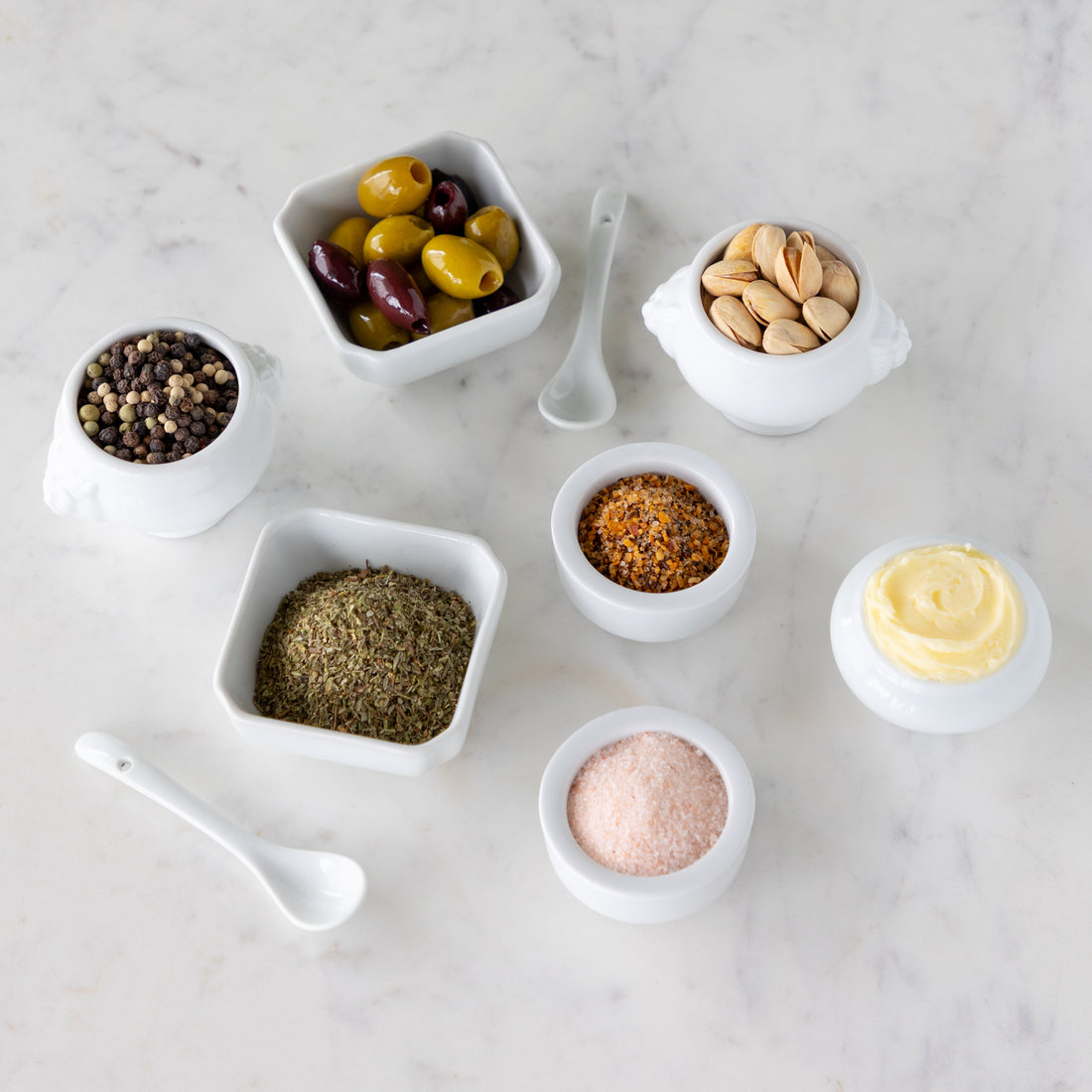 Assorted spices and seasonings in small white bowls next to white spoons on a marble surface.
