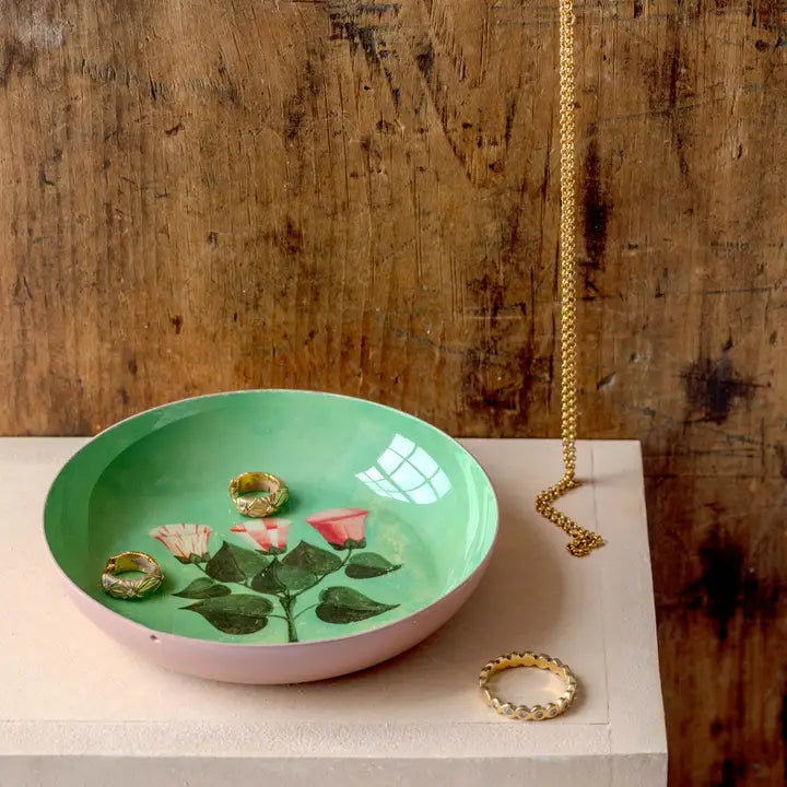 A green enamel plate with illustrated pink florals holding gold jewelry on a table.