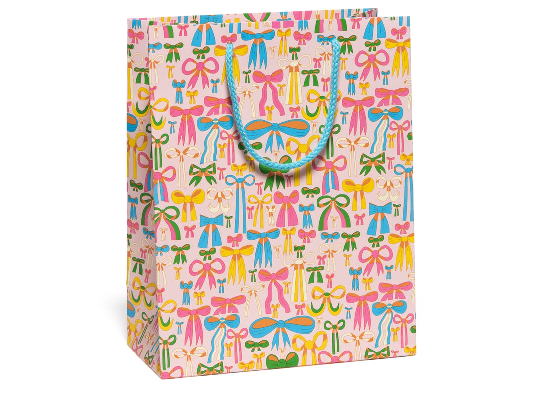 Gift bag with illustrations of lots of bows in multiple colors and styles all over it with a pink background and a bright blue handle. 