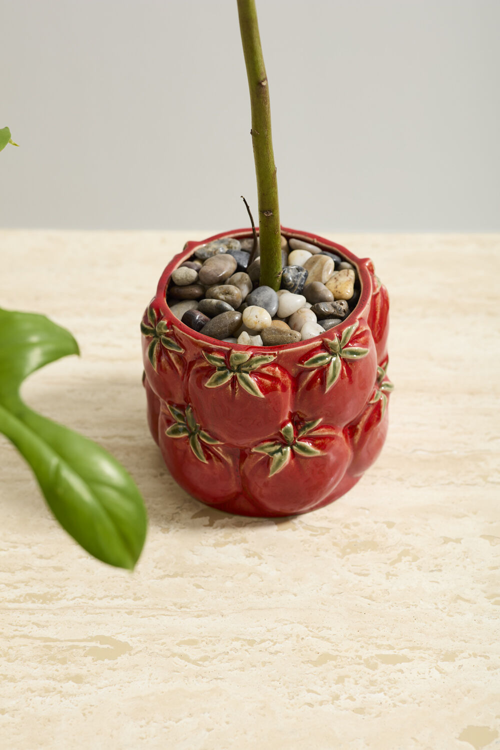 Red decorative pot with pebbles and a plant on a light surface.