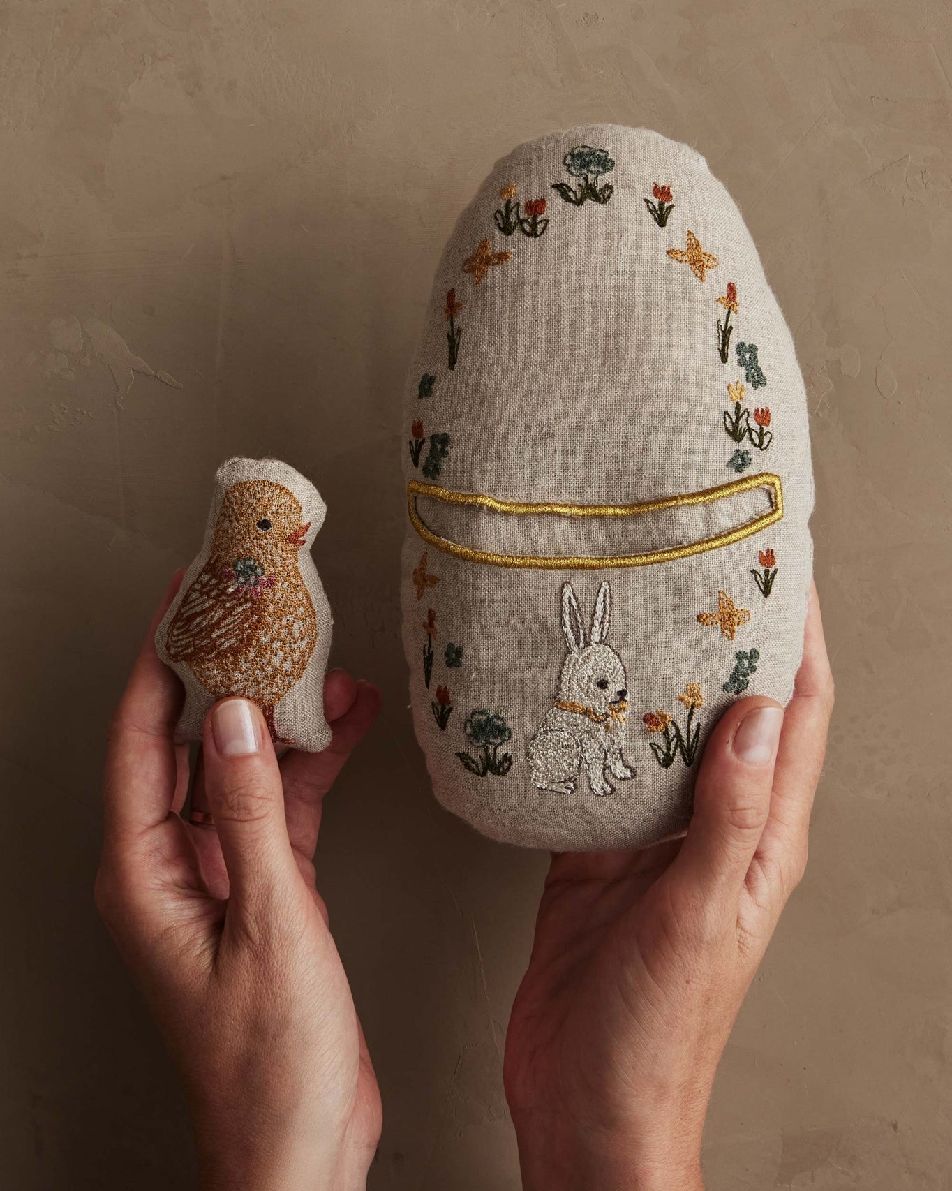 Embroidered fabric egg with a rabbit design held by hands against a beige background.