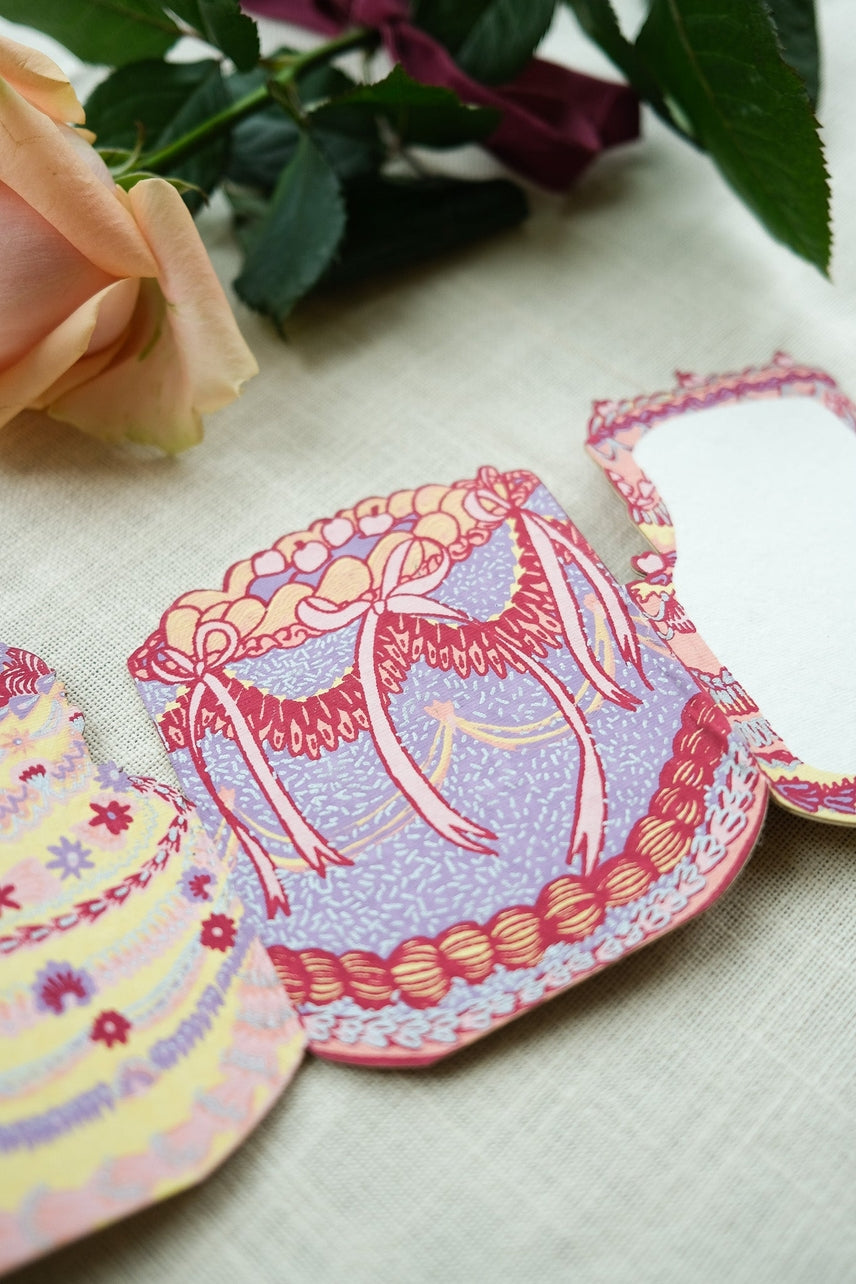 Decorative paper card of a birthday cake on a textured surface with a flower in the background.