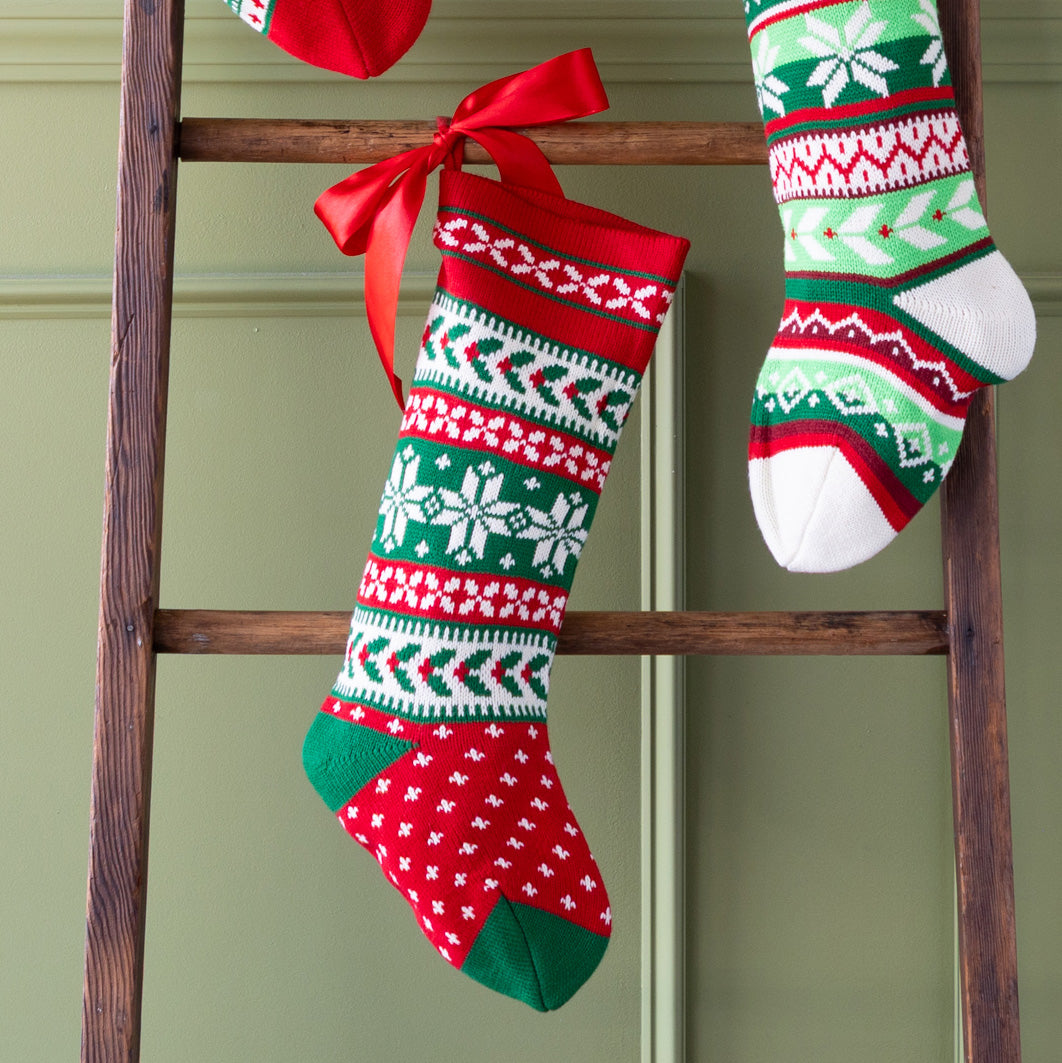 Decorative Christmas stockings with red, green, and white patterns hanging on a wooden ladder against a green wall.