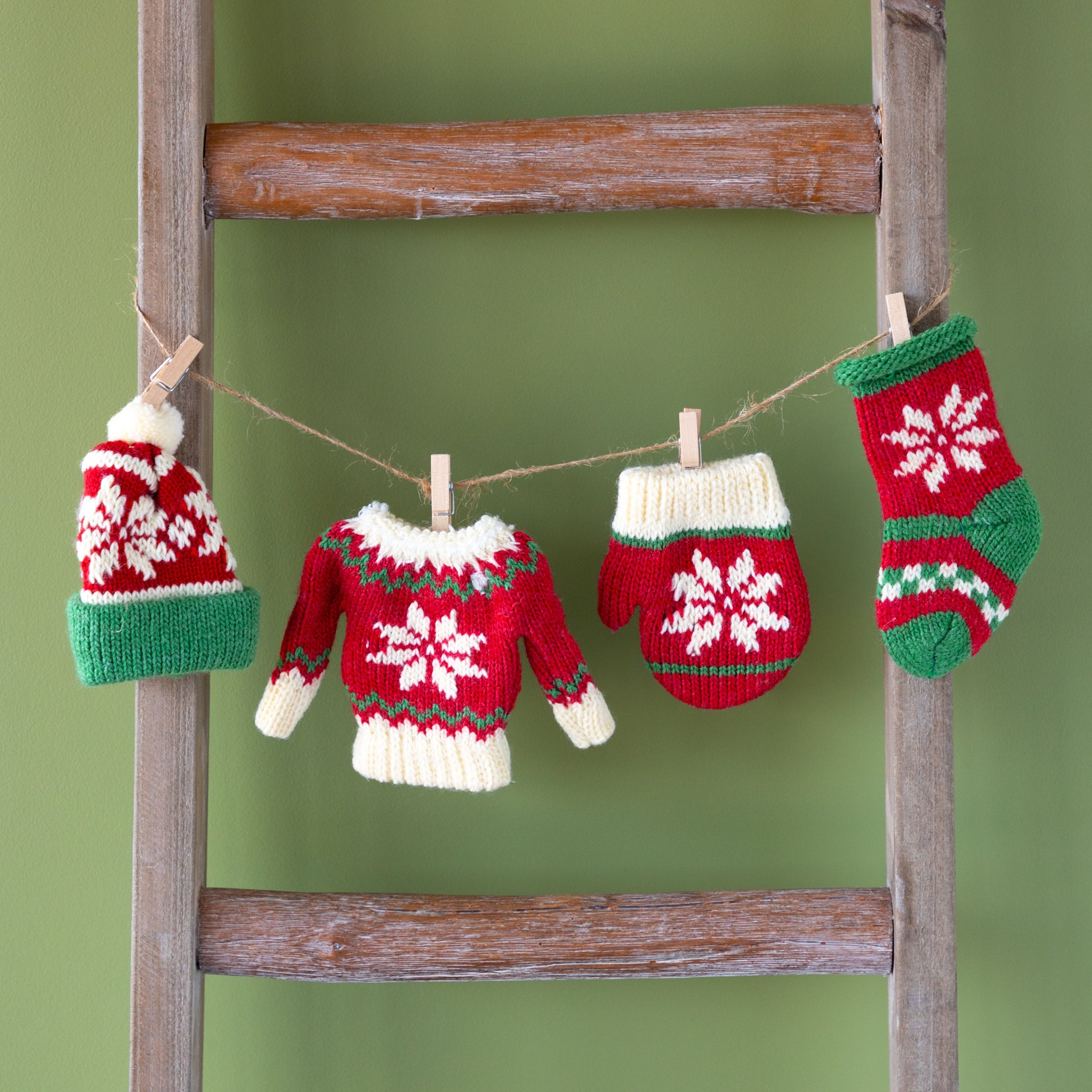 Knitted Christmas-themed items including a hat, sweater, mitten, and stocking hanging on a wooden ladder against a green background.