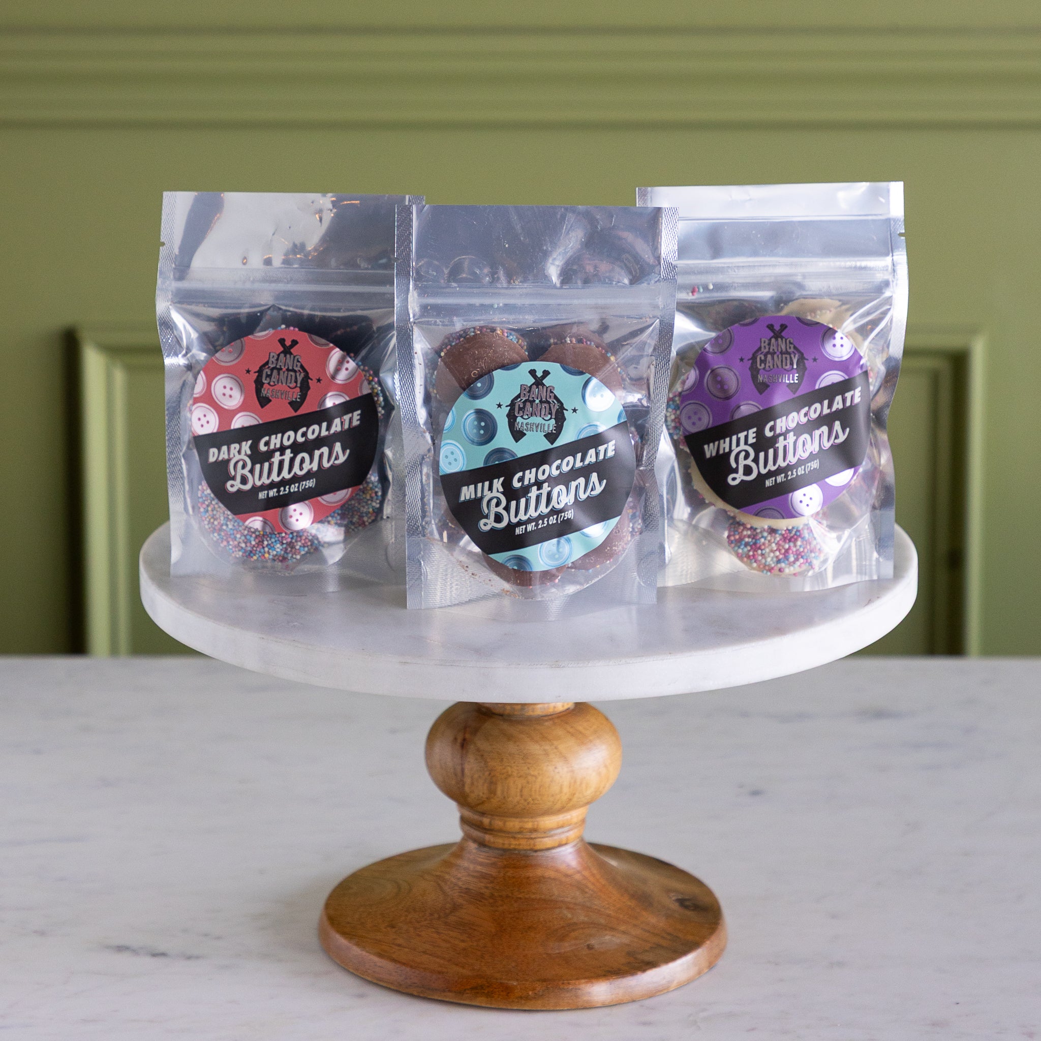 Three packages of chocolate buttons on a wooden stand with a green wall background.
