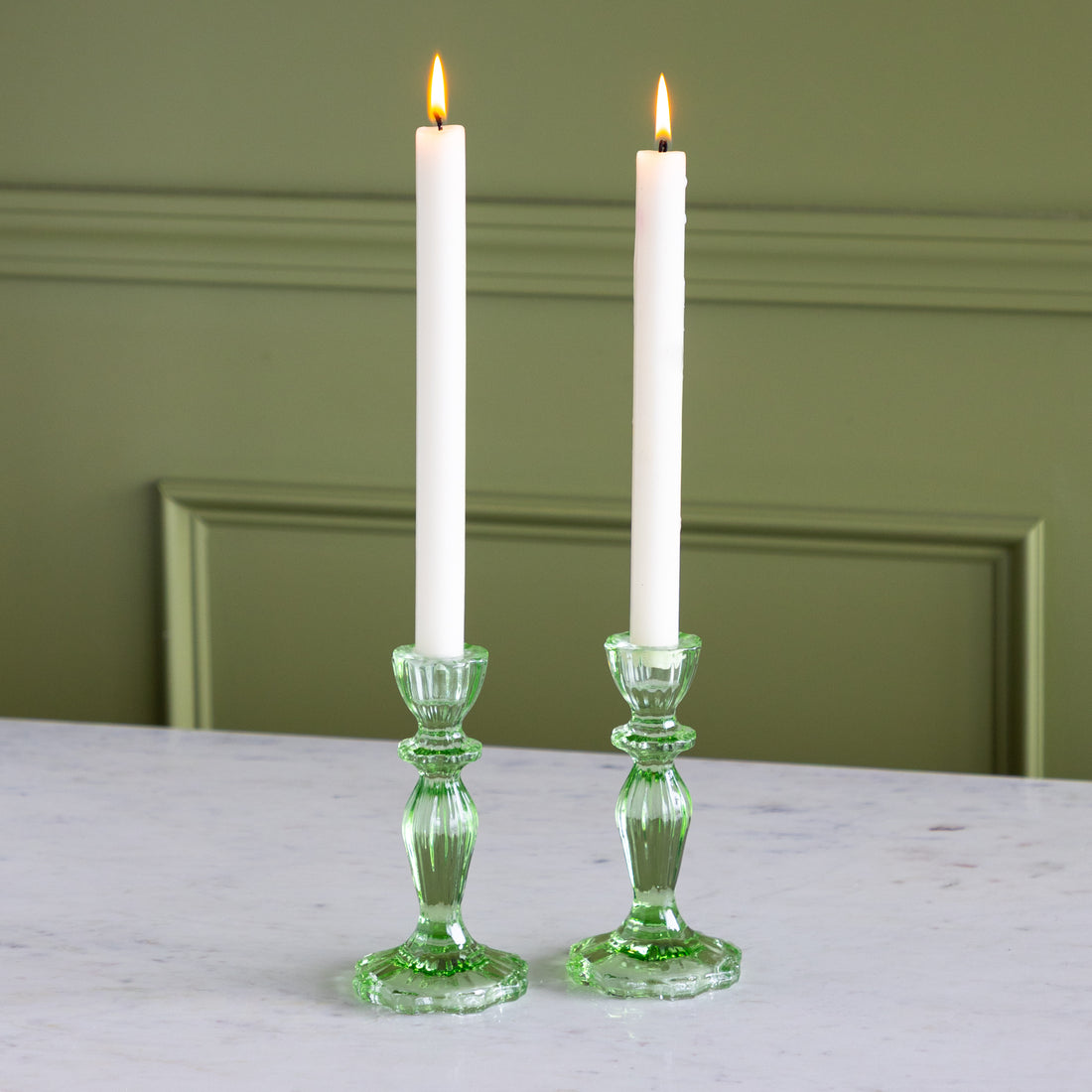 Two white candles in green candlesticks on a marble surface with a green wall background.
