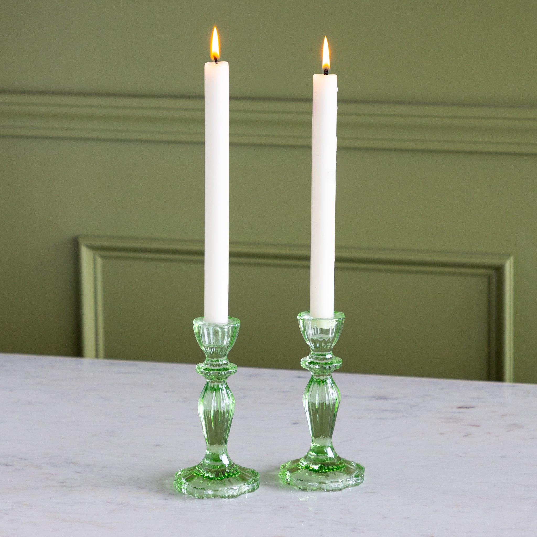 Two white candles in green candlesticks on a marble surface with a green wall background.