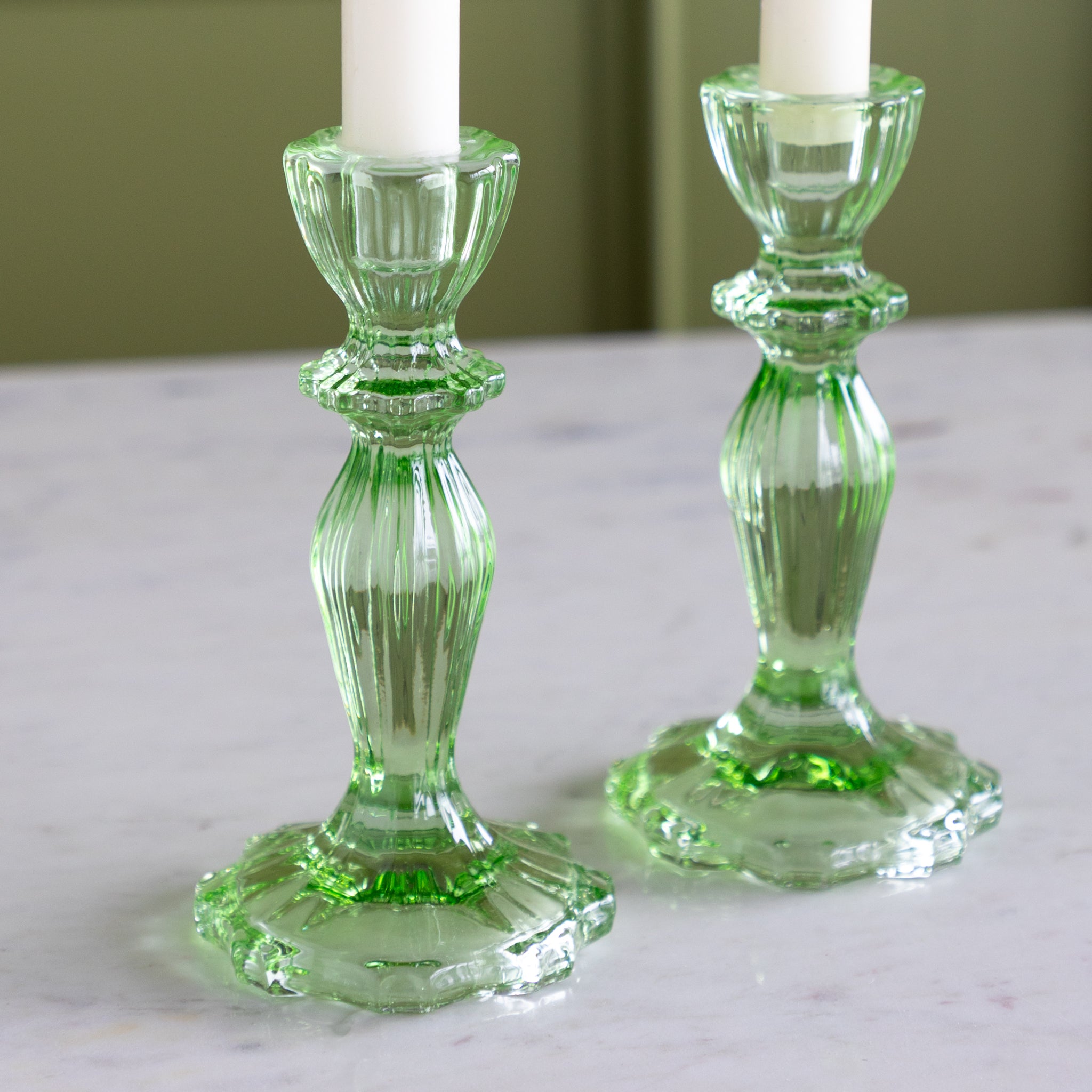 Two green glass candle holders with candles on a marble surface.