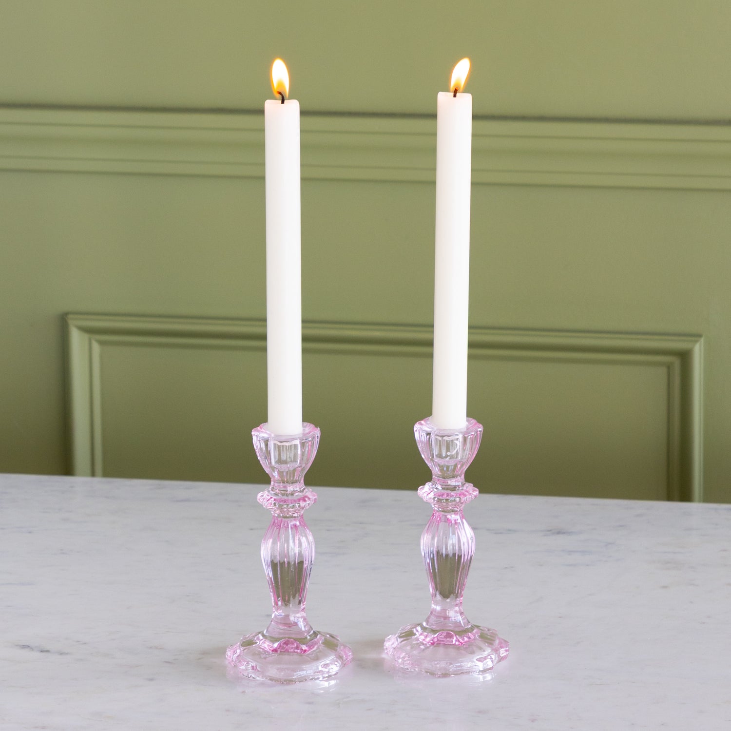 Two lit candles in pink glass candle holders on a white surface with a green wall background.