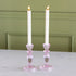 Two lit candles in pink glass candle holders on a white surface with a green wall background.