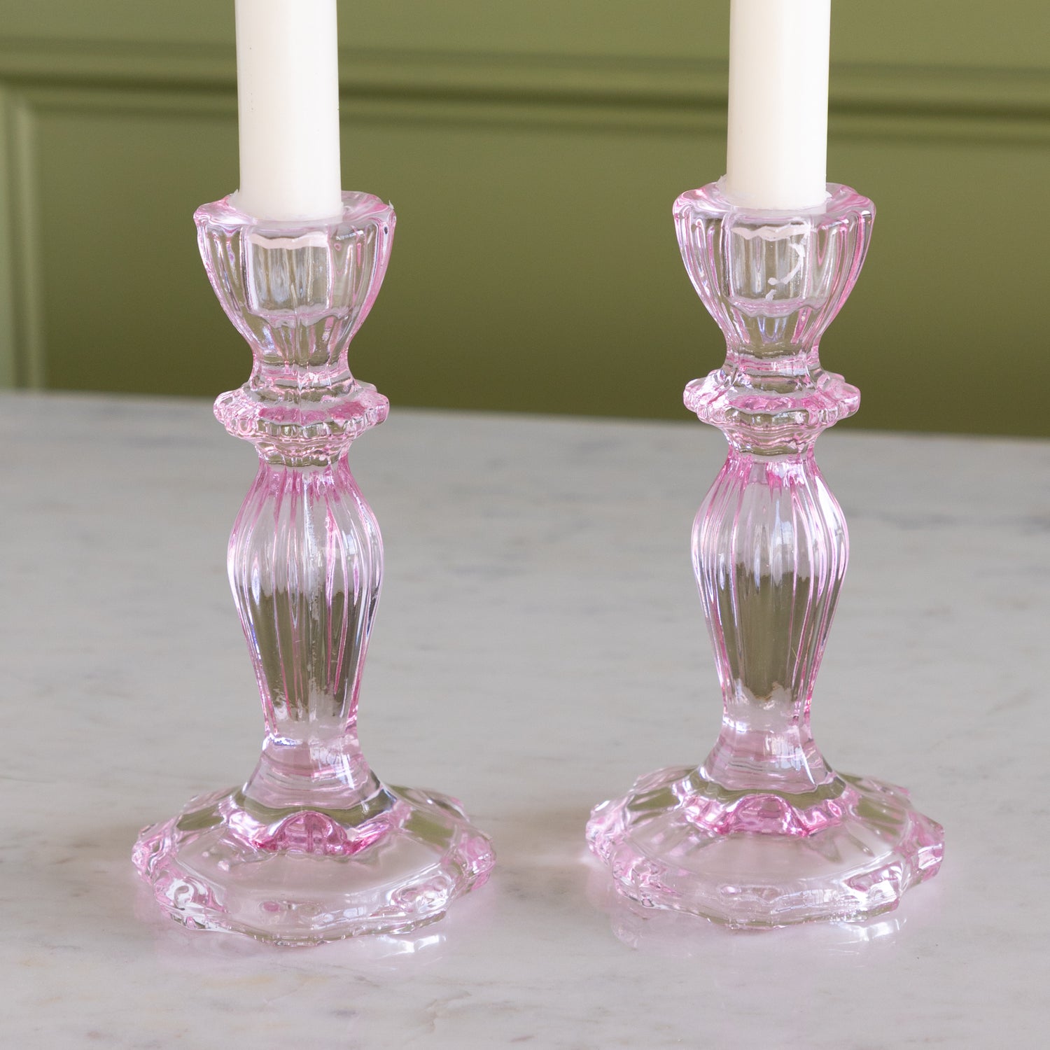 Two pink glass candlestick holders with white candles on a marble surface.