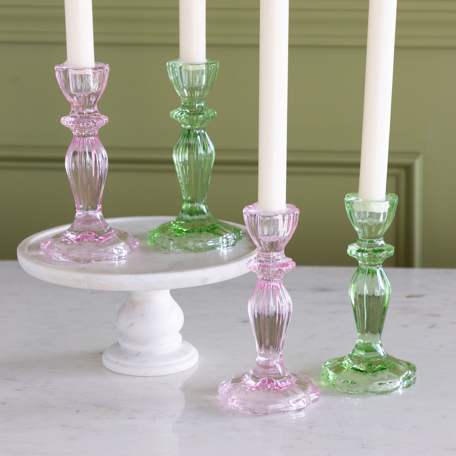 Four glass candlesticks with pink and green bases on a white surface against a green wall.