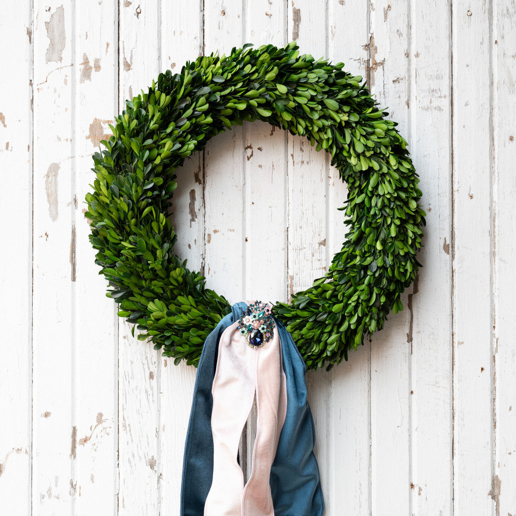 A Mills Floral Boxwood Wreath hanging on a white wooden wall, with a decorative broach and ribbons.