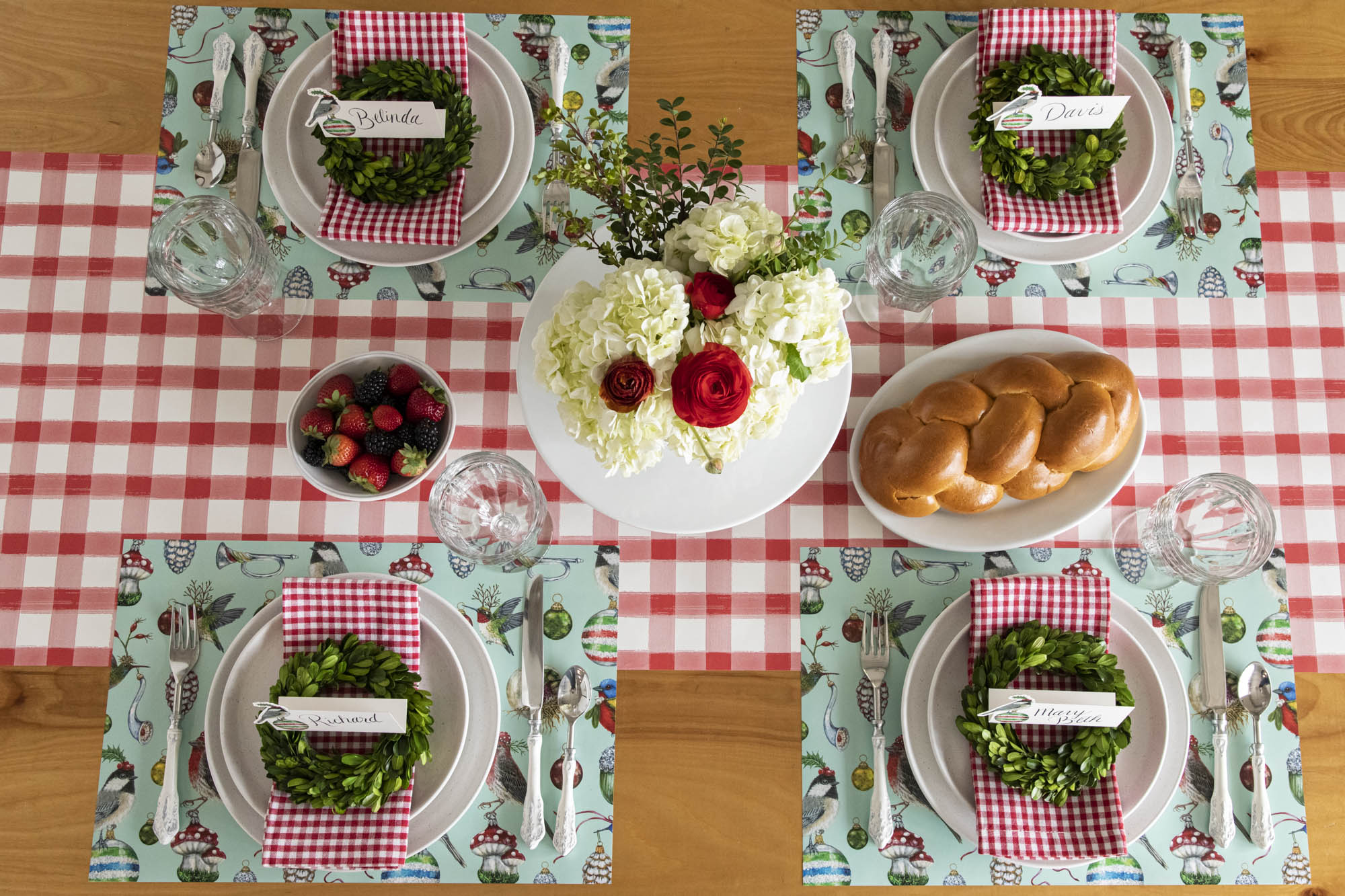 A Christmas table setting featuring a floral centerpiece, the Baubles & Birds Placemats, boxwood wreaths, Christmas Chickadee Place Card and red-and-white checkered napkins on the Red Painted Check Runner.