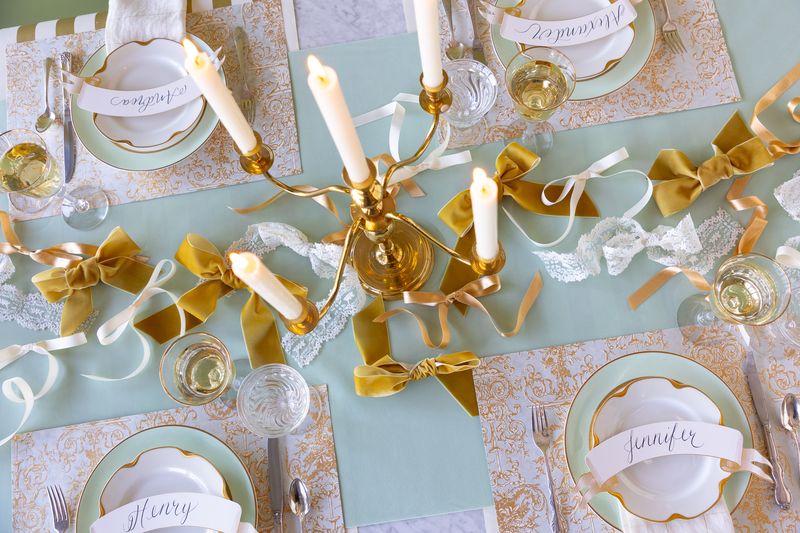 Decorative table setting with gold candles, ribbons, and place cards on a light blue tablecloth.