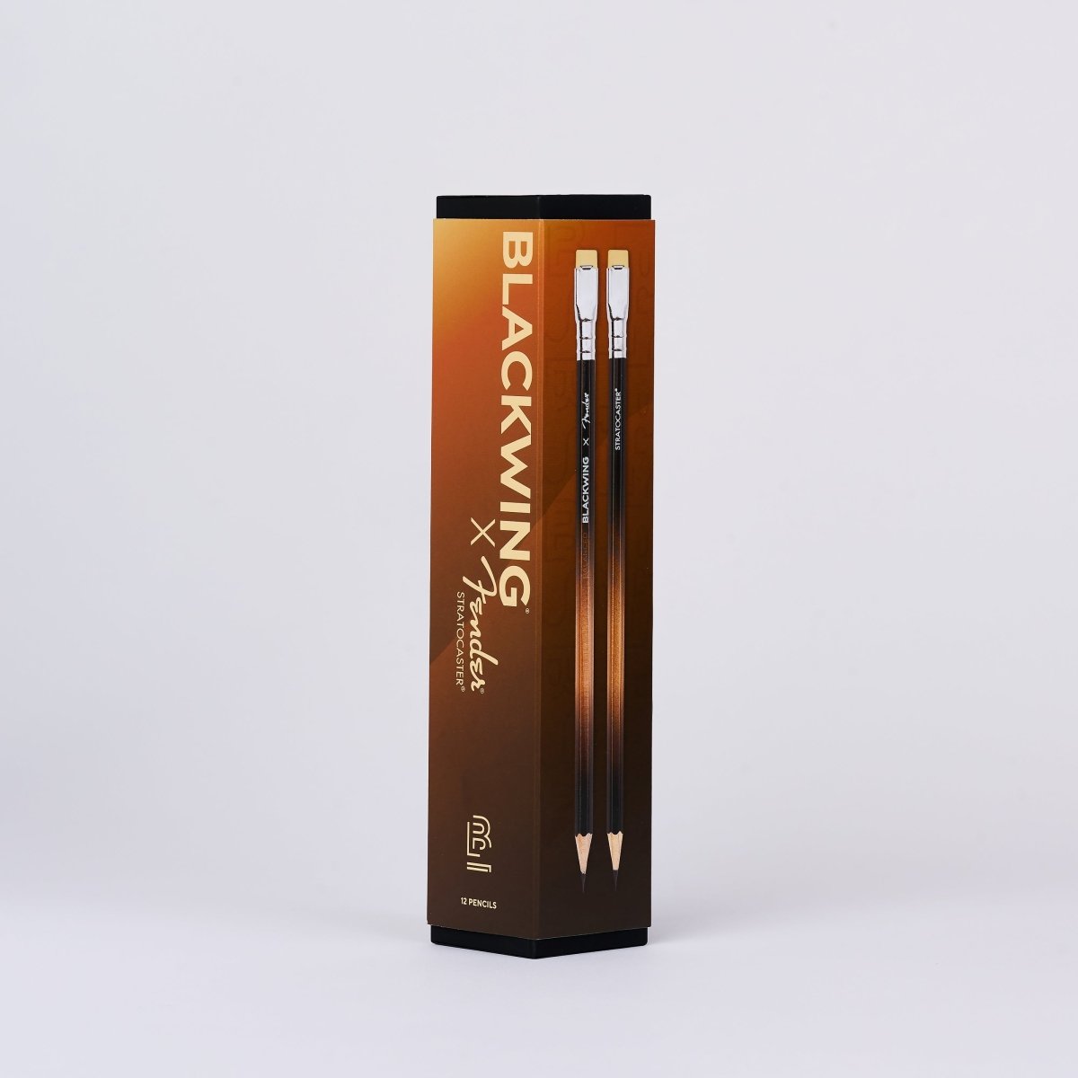 A tall rectangular product box for  Blackwing x Fender pencils, standing upright on a light gray studio background. The box is dark brown and black with gold gradient text and shows two pencils inside.