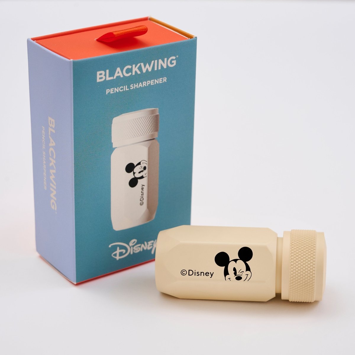 A Disney Blackwing pencil sharpener shown next to its blue and orange retail box. The sharpener itself is a light beige/cream color with a black Mickey Mouse head logo and &