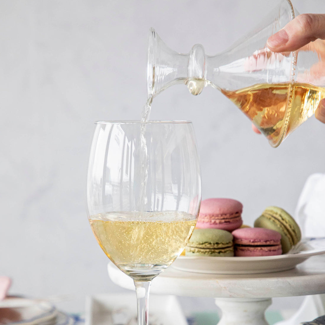 A person pouring white wine into a glass with the Mouth Blown Clear Vase 
 featuring macarons on a plate sitting on a riser in the back.