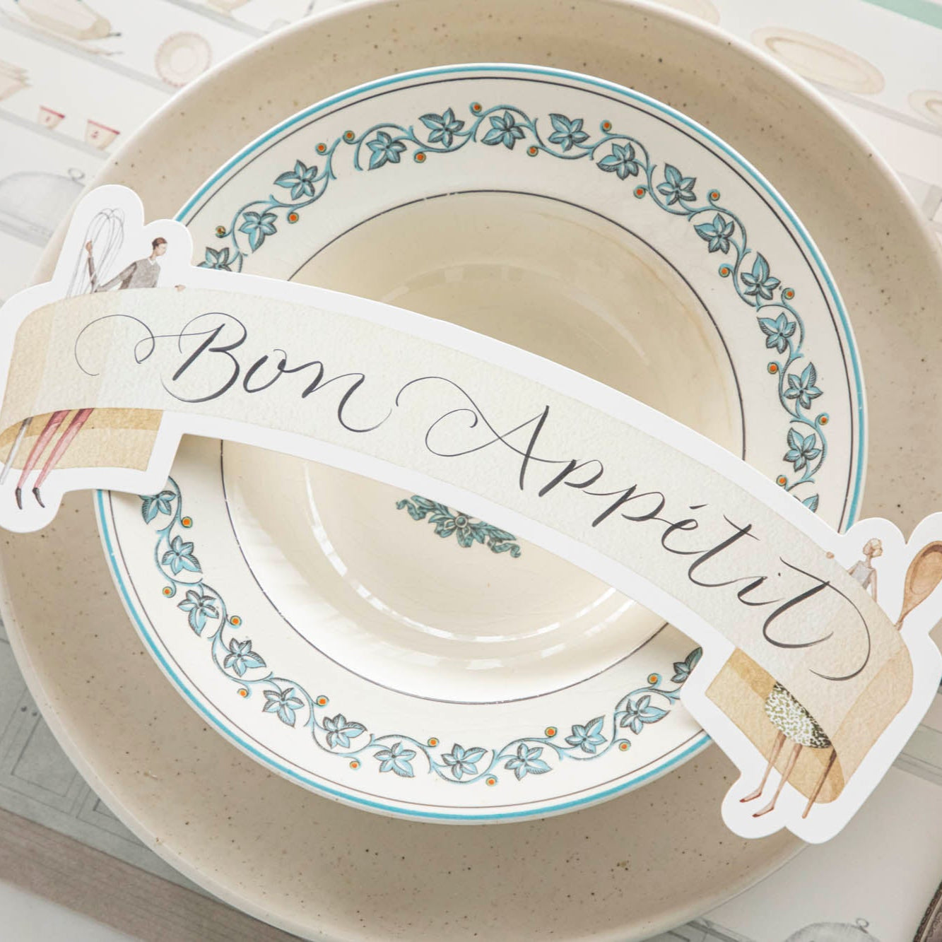 Two Cooks Banner Table Accent with &quot;Bon Appetit&quot; written on it, resting on dinner plates, on an elegant place setting for one.